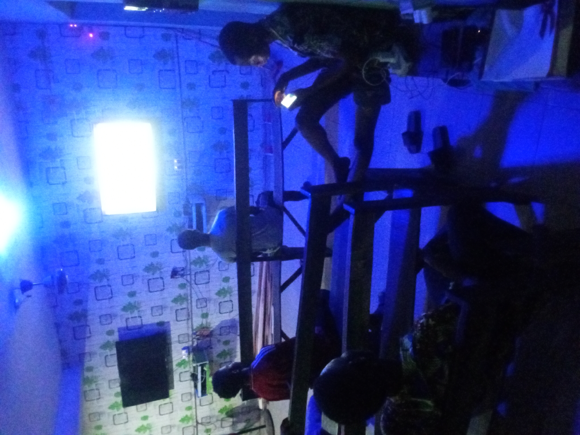 The interior scene, taken in Unknown, Unknown, is illuminated by a bright rectangular light source on a wallpapered wall with a repeating square and floral pattern. A male figure is positioned on a dark, wooden structure, possibly a ladder or scaffolding, and is looking down at a brightly lit mobile phone in his hand. Various other blurred figures are present around the structure. The lighting is primarily blue, casting heavy shadows and obscuring details. Other objects visible include a dark rectangle on the wall, potentially a TV screen, and a fan.