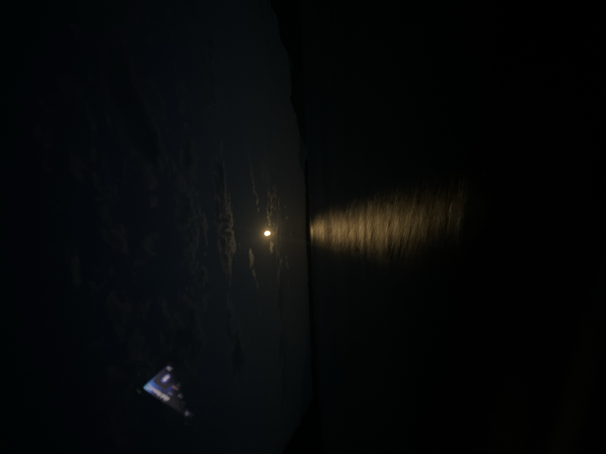 The image shows a dark night scene, possibly taken near the city of Vranjina, Montenegro, given the context. The left side of the image displays a dimly lit body of water or lake, possibly Lake Skadar. A small, faint light source reflects on the water. To the right, a dark wall or structure divides the scene. A blurry light streak is cast onto the black wall, possibly from a moving vehicle. In the lower-left corner, a lit electronic device, possibly a smartphone or tablet, is faintly visible. No people or specific subjects are clearly discernable. The environment appears to be rural or near a body of water.