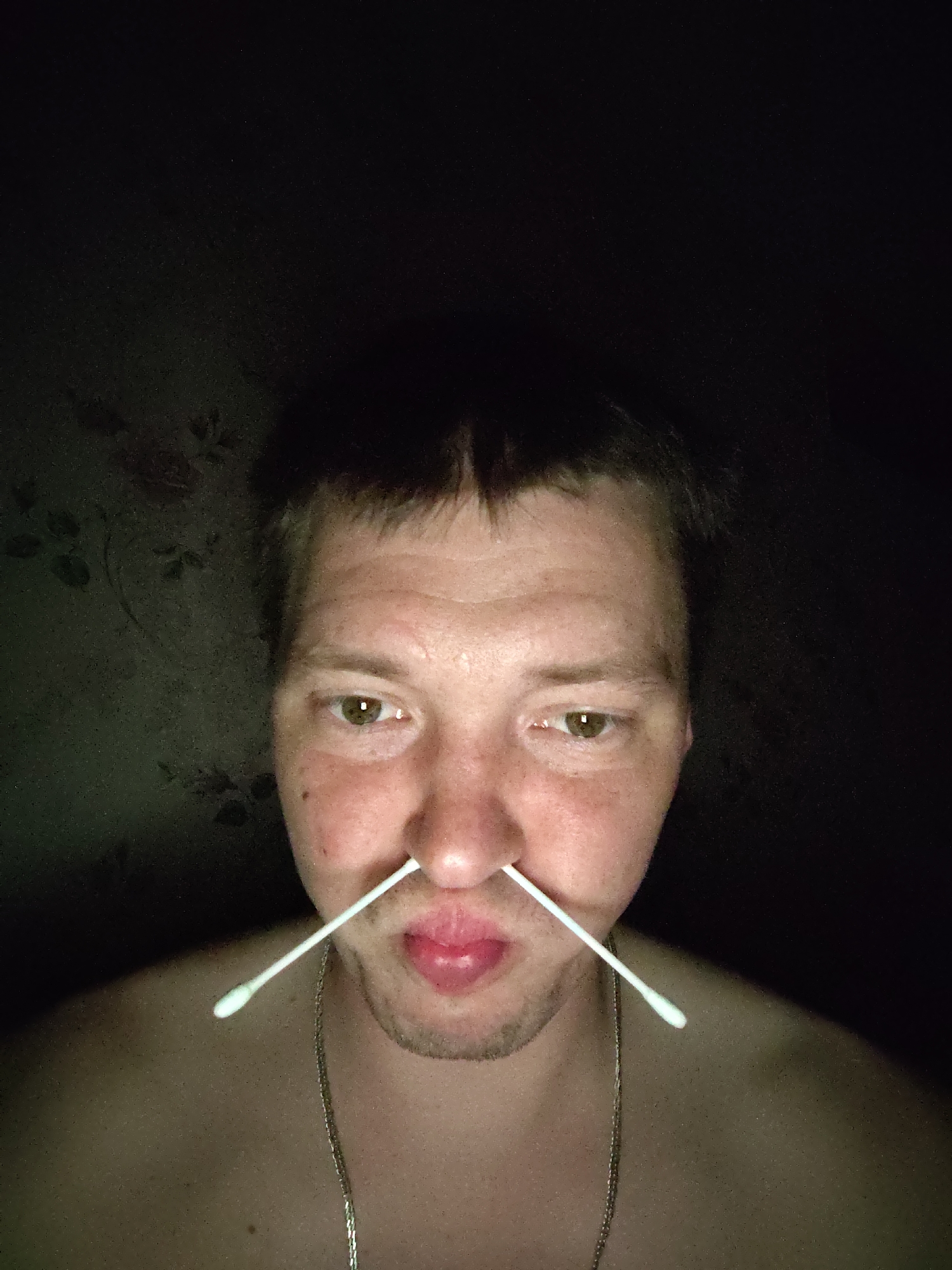 A head-and-shoulders shot of a man with fair skin and short brown hair. Two cotton swabs are inserted into his nostrils, pointing outward. He is wearing a silver chain necklace. The background consists of a dark wall with faint floral patterns on the left side and solid dark areas on the right. The location is likely indoors, somewhere in the city of Марковский, Russia.