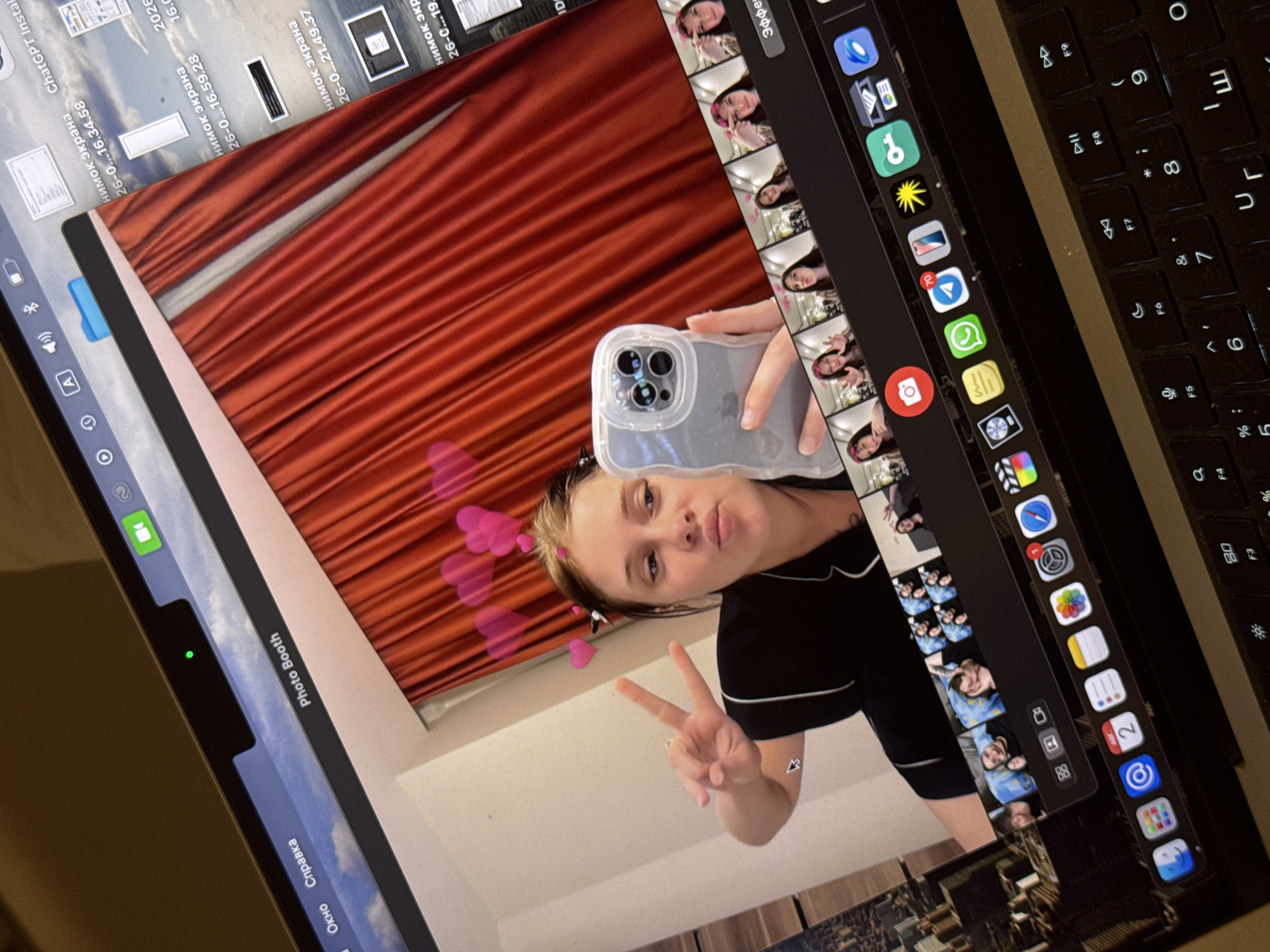 A young woman is taking a selfie in a room with red curtains in the background. She is wearing black pajamas and has pink hearts floating around her head. She is holding up her phone and making a peace sign with her fingers. The selfie is being taken on a laptop computer, which displays the "Photo Booth" application. The laptop is open on a surface, and a keyboard is visible in the foreground. The environment is indoors. There are no visible indicators linking this image to Aksu, Turkey.