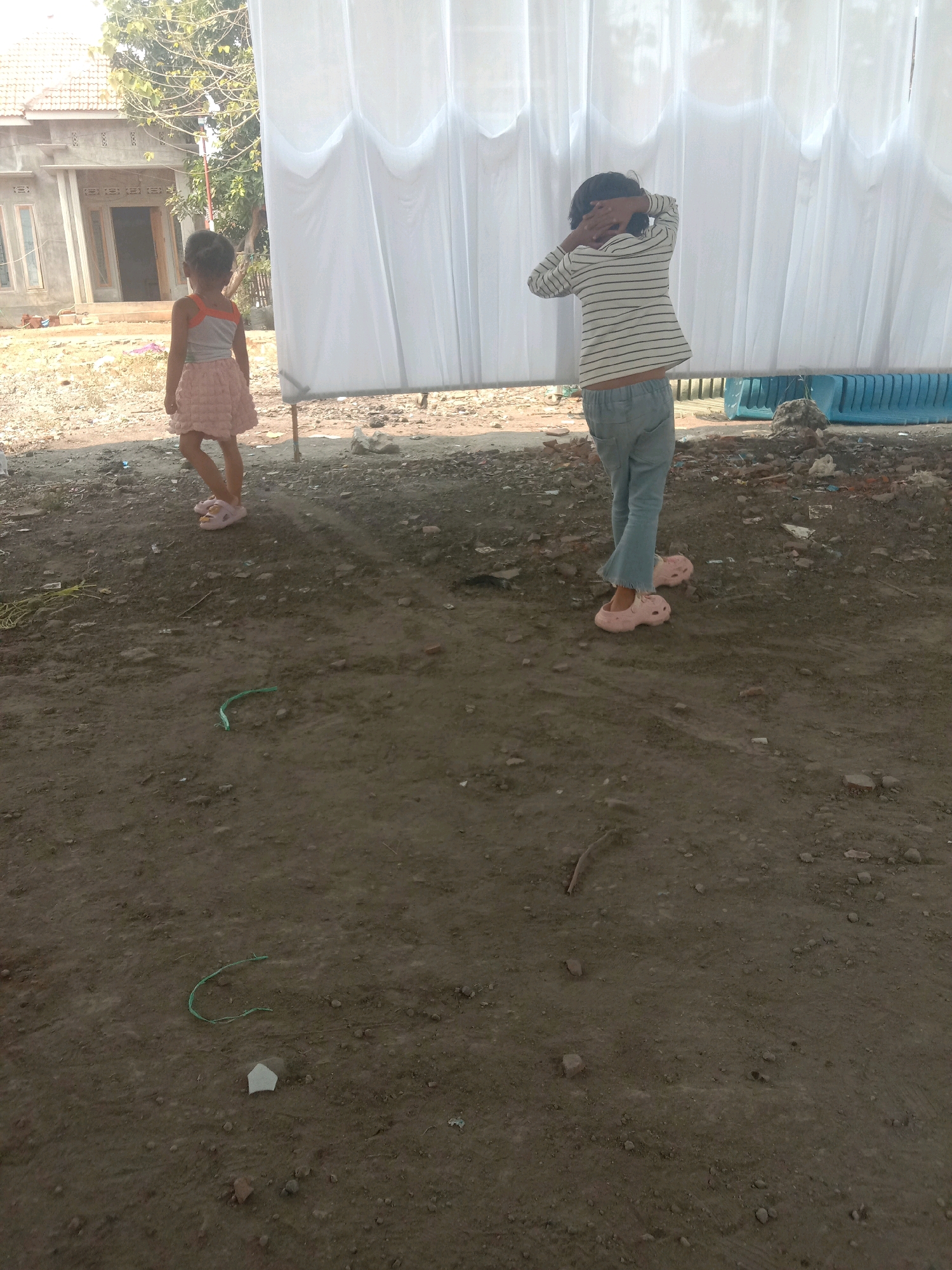 This image depicts an outdoor scene in Karang Anyar, Indonesia, characterized by a dirt ground with scattered debris. Two young girls are present. The girl on the left, wearing a pink skirt and a sleeveless top, stands facing away from the camera. The girl on the right, dressed in a striped long-sleeved shirt and light-colored pants, has her hands behind her head, obscuring her face from view. Behind the girls, a large white curtain is suspended, and to the left, a building under construction is visible with its roof and entryway. The ground is uneven, with patches of dry dirt and some small plants or weeds.