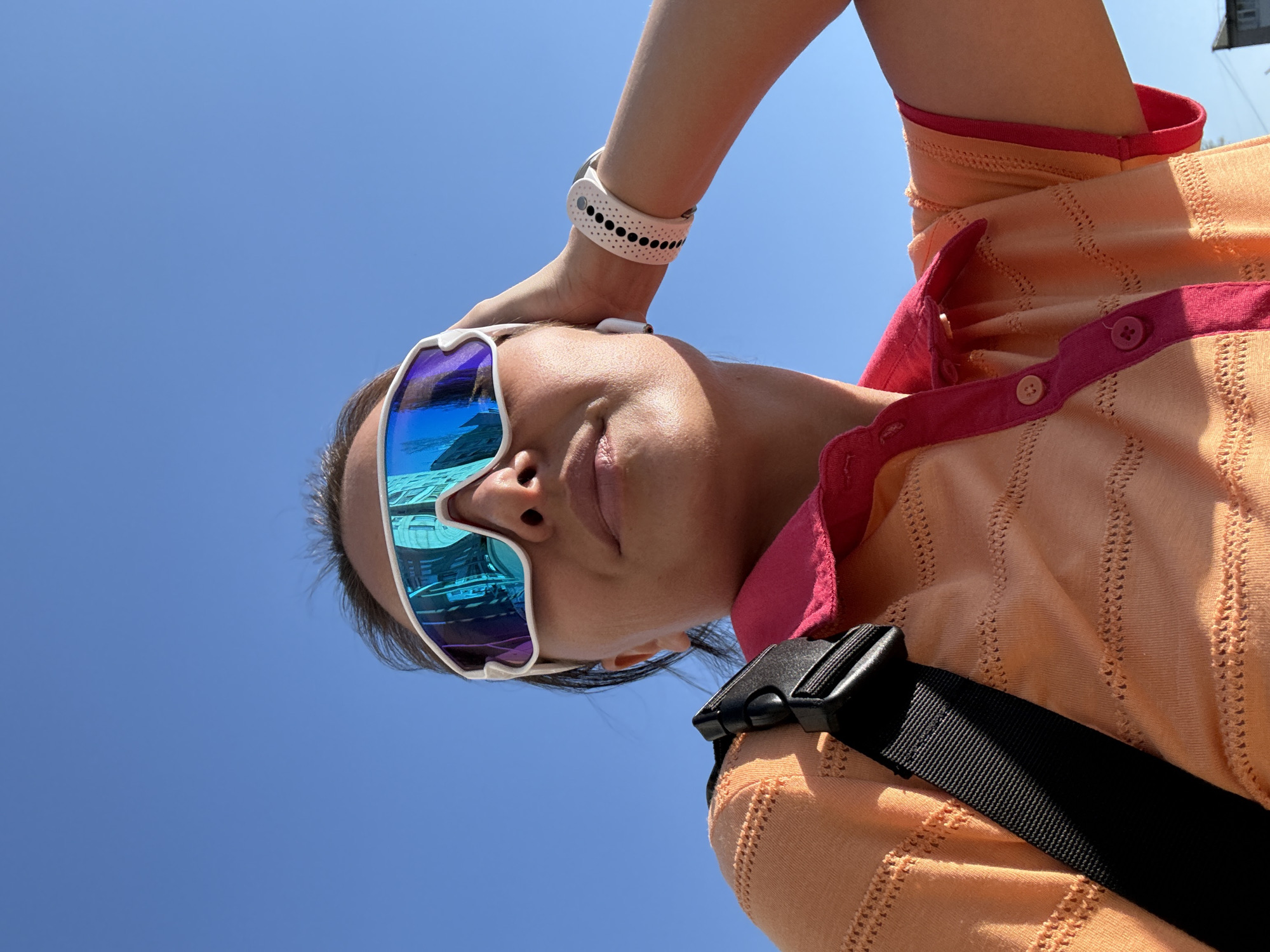 A low-angle shot depicts a woman wearing mirrored sunglasses, an orange polo shirt with pink trim, and a white wristband, set against a clear blue sky. Her head is tilted back, and her arm is raised to her forehead. A black strap crosses her shoulder. The reflection in the sunglasses appears to show a building. While the city of Chaikovsky, Russia, is mentioned, specific landmarks or context within the image that confirms this location are not visible.