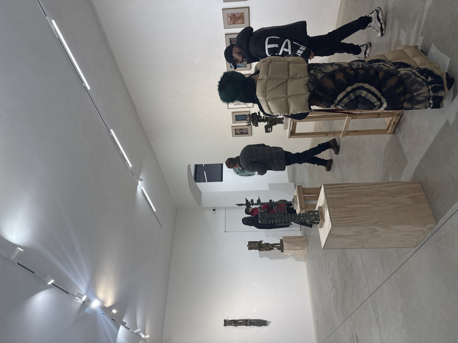 An art gallery displays diverse sculptures on pedestals. Two individuals, one in an Emporio Armani jacket and another in a puffy vest, observe artwork mounted on wooden structures, while another visitor stands nearby. Linear lights illuminate the white walls. In the background, a doorway provides a view into another room.