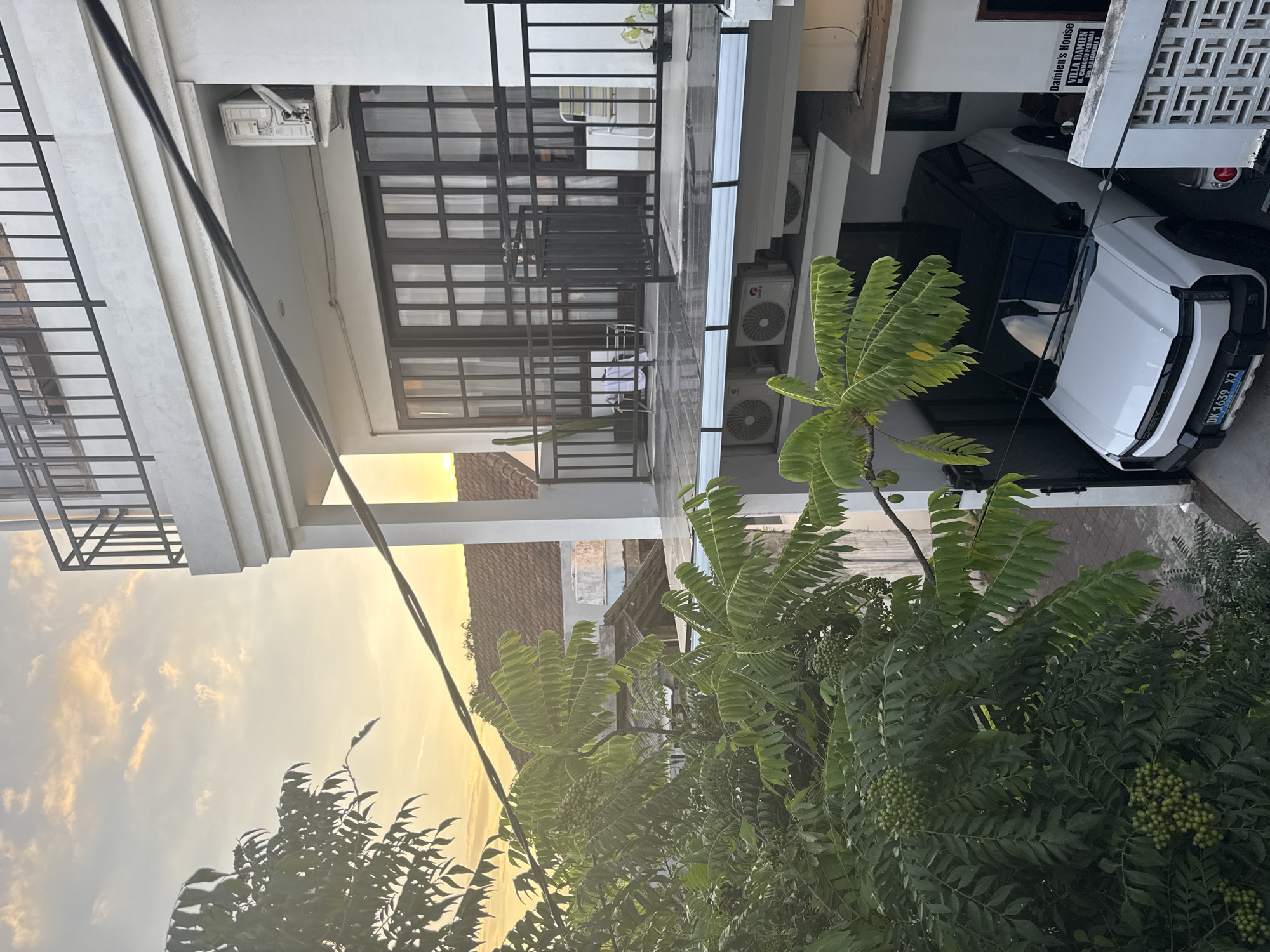 The image captures a two-story building with a white exterior and dark-framed windows, possibly located in North Kuta, Indonesia, as suggested by the license plate on the white car parked in the carport on the right. The building features a balcony with a black metal railing. Air conditioning units are visible on the side of the building. Lush green foliage fills the foreground. A power line stretches across the sky, which is tinged with the colors of sunset. A sign on the building identifies it as "Damien's House, Villa Damien" located on JL. Canggu Permai.