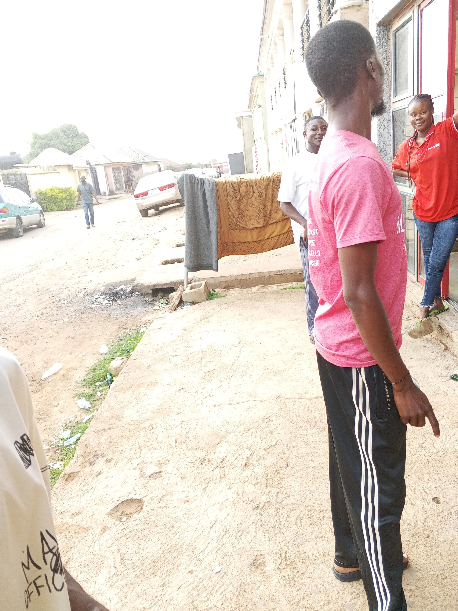 A street scene in Jos, Nigeria, captures several individuals and their surroundings under bright daylight. In the foreground, facing away from the viewer and slightly to the right, is a man with short dark hair and a partial beard, wearing a pink t-shirt and black track pants with white stripes down the sides. His left hand is visible, pointing downwards. The track pants have "IONAL ARTS CLUB" printed on the side. Partially visible text on his t-shirt includes "EAST," "CELLO," and "ANCHE."
To the right, next to a building, a woman in a red polo shirt and blue jeans smiles while looking out of a doorway. She wears light green sandals. Standing between her and the man in pink is another man in a white t-shirt, also smiling and looking towards the camera.
The immediate ground is a mixture of concrete and dirt, with a visible drainage ditch containing burnt debris and litter. Further down the dirt road on the left, a man in a blue shirt and jeans walks away from the viewer. Two cars are parked or driving on the dirt road: a teal/green sedan on the left and a white sedan on the right. Clothes, including a brown patterned fabric and a grey piece, hang on a drying line or rail. In the background, various buildings are visible, some appearing residential, with one showing partial vertical text that reads "PHARMACY." The bright lighting suggests it is daytime with clear weather. A small portion of a light-colored shirt with the text "MA OFFICE" is visible in the bottom left corner.
