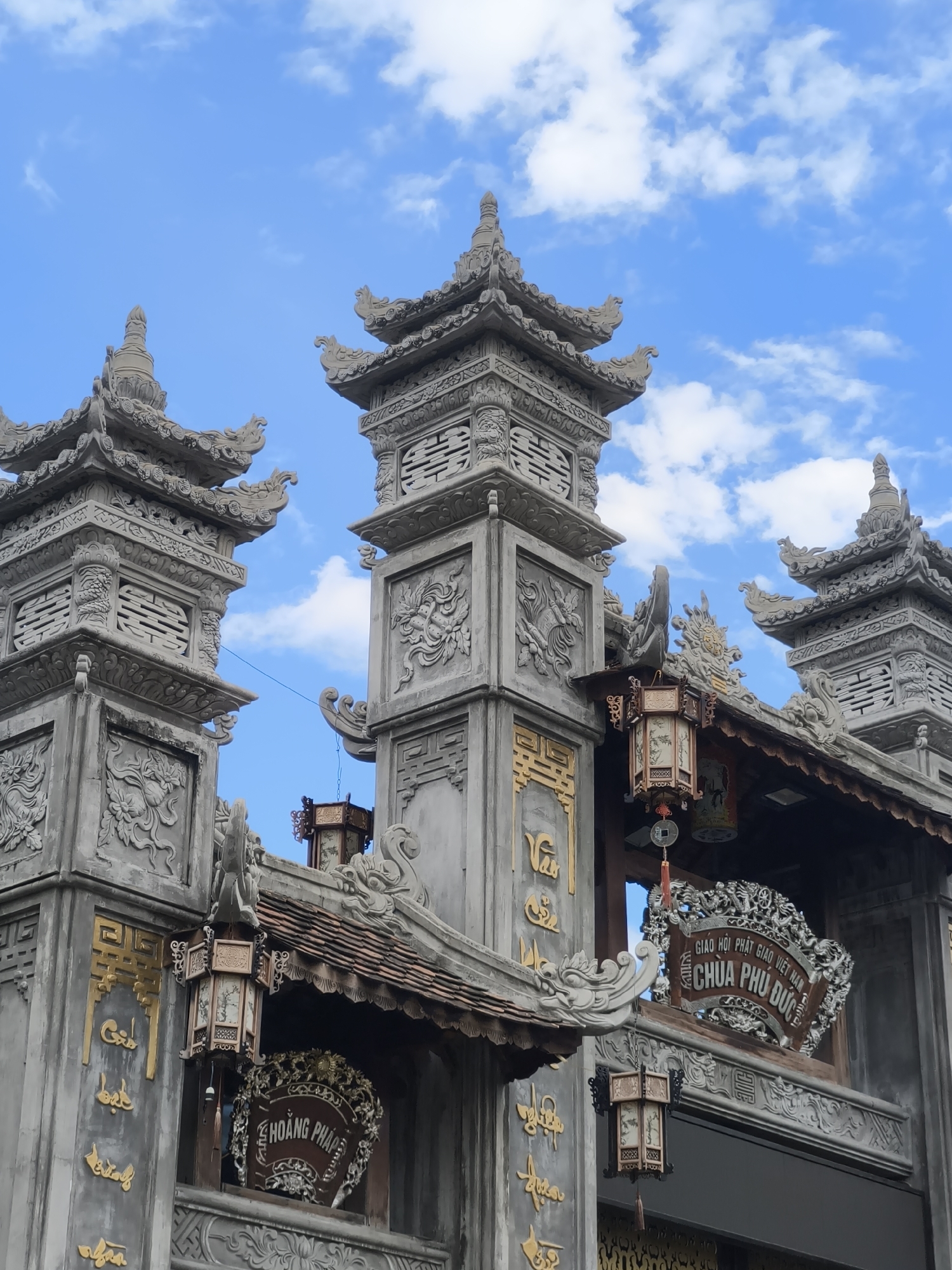 The image shows an ornate stone gate with traditional Vietnamese architectural elements. The gate is decorated with intricate carvings, including dragons and floral motifs. Several lanterns are hung from the roof of the gate, adding a warm glow to the scene. A sign with Vietnamese writing hangs prominently above an entrance. The gate is set against a backdrop of a bright blue sky with scattered white clouds. The overall impression is one of grandeur and cultural significance. This scene is located at Chua Phu Duc in Nha Trang, Vietnam.
