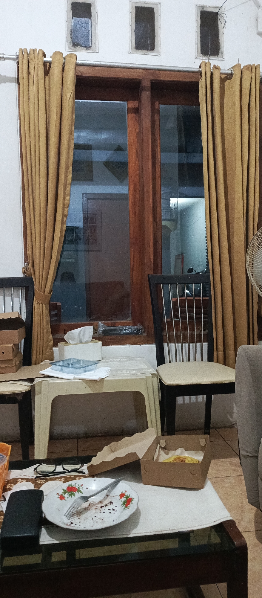 The image depicts an indoor scene, likely a dining area or living room, with a prominent window and curtains. The window has wooden frames and glass panes, revealing a nighttime view of an exterior area with a motorcycle and buildings. Two chairs are visible, one on the left with a black frame and dark seat, and another on the right with a black frame and a light-colored seat. In front of the window, a small, light-colored plastic table holds a tissue box and a clear plastic container with dividers. To the left of the table, stacked cardboard boxes are visible. A larger coffee table in the foreground displays a plate with remnants of food and a fork, a black rectangular object, a pair of eyeglasses, and an open cardboard box containing a food item. The room appears to be located in Situbondo, Indonesia, as indicated by the context.