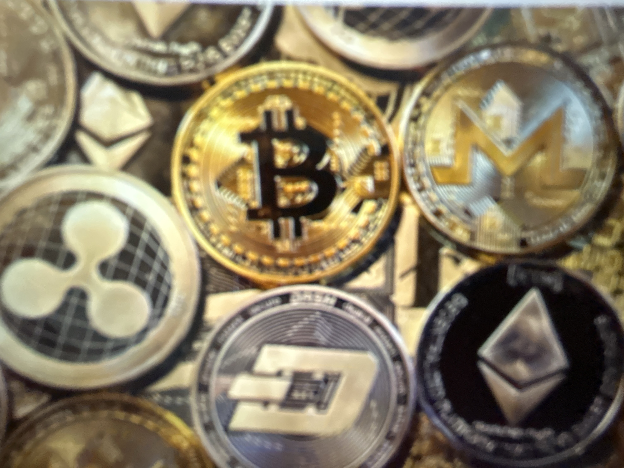 The image displays a close-up, top-down view of several cryptocurrency coins arranged on a textured surface. The coins are varied in color, including gold, silver, and black, and feature distinct logos. Prominently visible are coins with the Bitcoin symbol (a stylized "B"), the Ripple logo (three interlocking circles), the Dash logo (a stylized "D" within a circle), and what appears to be the Ethereum logo (a diamond shape). Other coins are partially visible and out of focus. The background is indistinct but appears to be a continuation of the textured surface. The image is not detailed enough to provide information about a specific location in Gelendzhik, Russia, or any people, actions, or interactions.