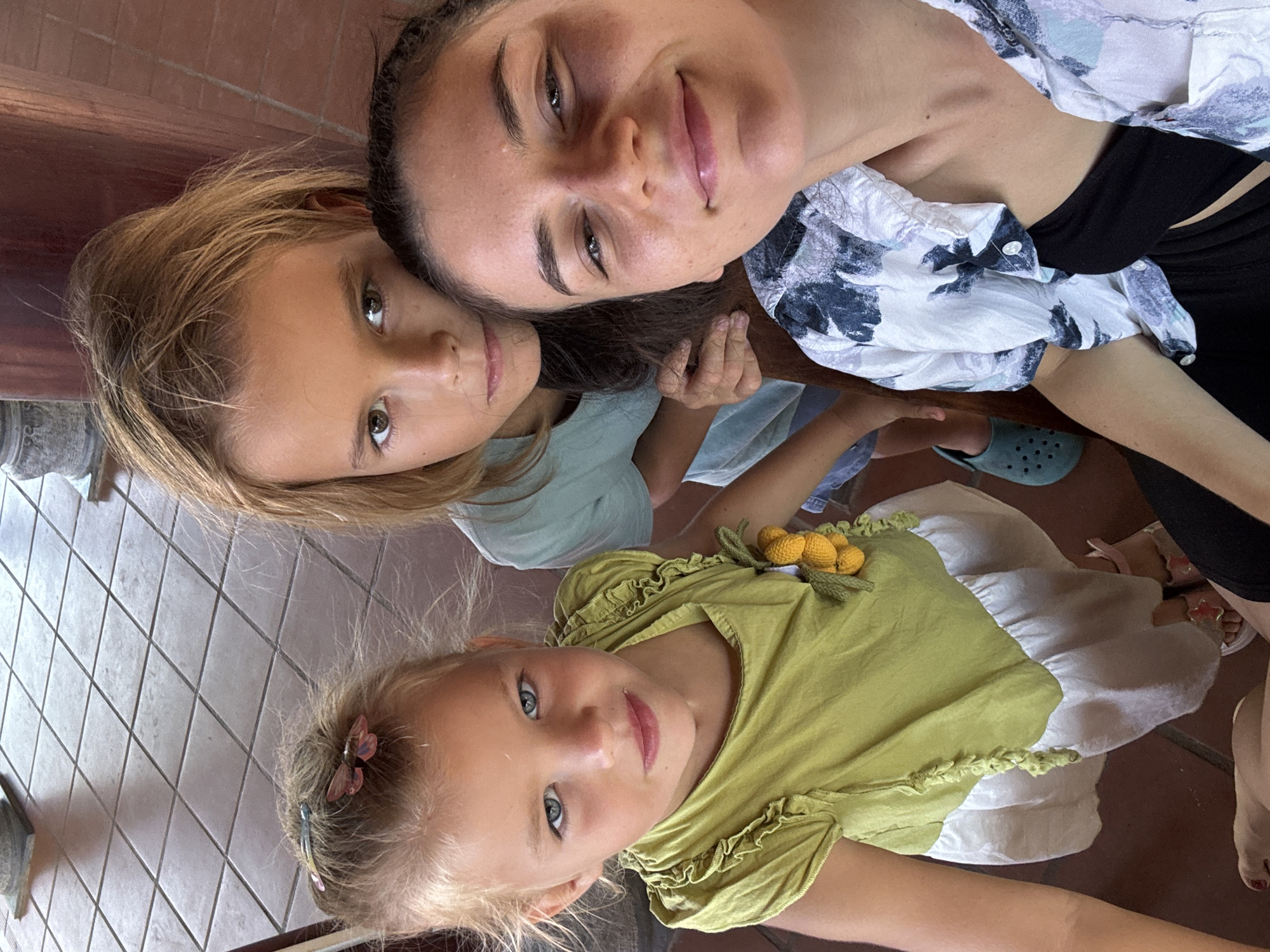 The image shows a selfie taken from above, featuring a woman and two young girls. The woman is wearing a white and blue floral-patterned shirt over a black top. One girl has blonde hair and is wearing a light green top. The other girl has blonde hair with a butterfly hair clip and is wearing a green dress with a lemon detail. The location appears to be indoors, possibly a restaurant or cafe, with a tiled floor. It is reported to be Mũi Né, Vietnam, but there is no visual information in the image to confirm this.