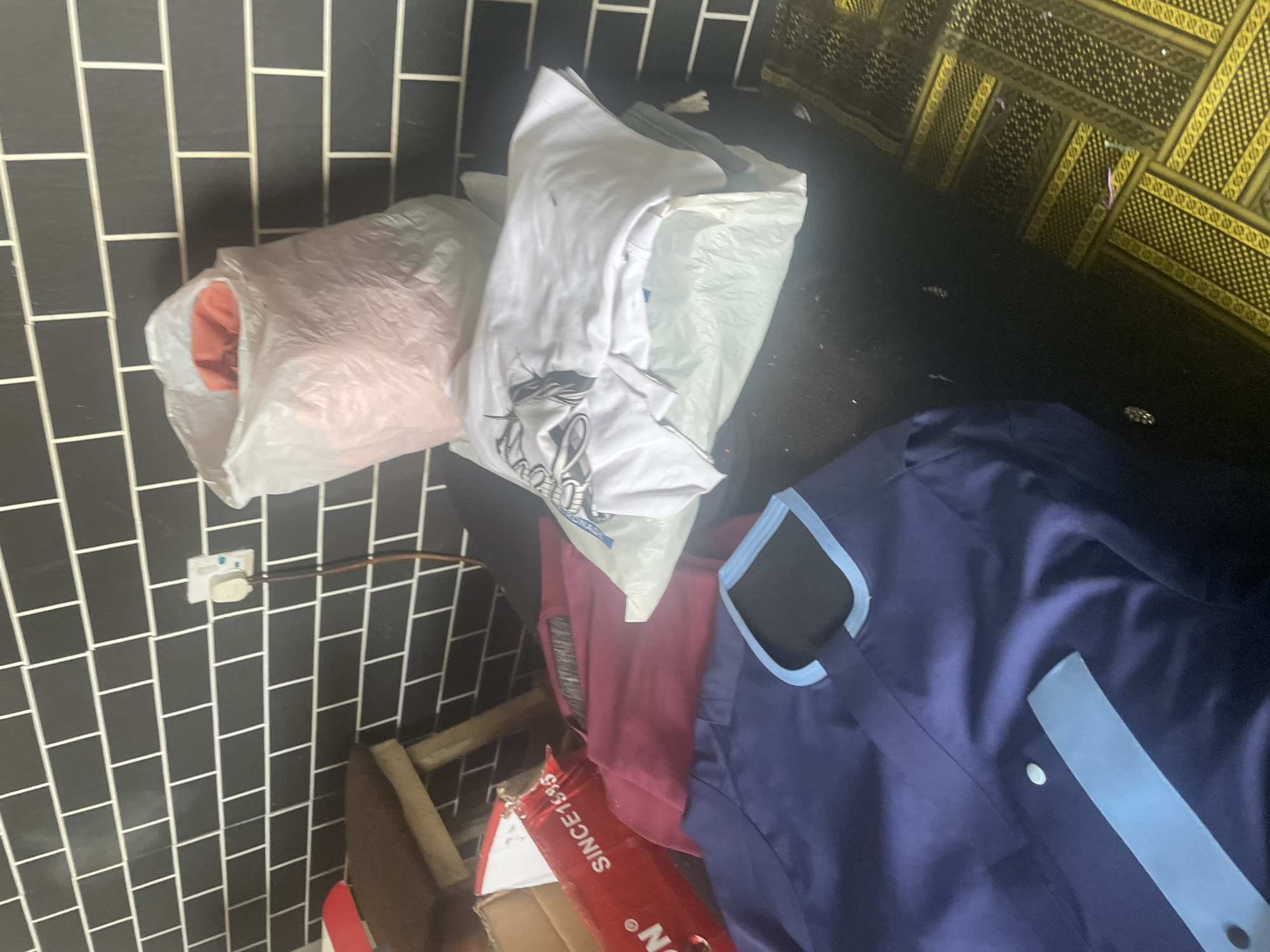 The image depicts a collection of clothing items placed on a dark surface, possibly a table or floor. The background features a black tiled wall with white grout lines, and a decorative yellow and black patterned fabric is visible in the upper right portion of the frame.
Objects in the foreground include several pieces of clothing. On the left, a light-colored plastic bag contains an orange garment. Next to it, another plastic bag holds a white t-shirt with a graphic design. A maroon-colored garment is situated below these, and to the right, a dark blue garment with light blue trim is prominent, appearing to be a traditional African attire, possibly a kaftan or agbada. A red box with white text is also visible near the bottom, with the partially readable words "SINCE1985" and "ON" visible.
The lighting suggests an indoor setting, possibly during the daytime due to the brightness from an unseen light source. No people are present in the image. The scene is cluttered, with clothing items haphazardly arranged. The location context of Jauro Yinu, Nigeria, suggests the blue garment might be traditional attire worn in that region.