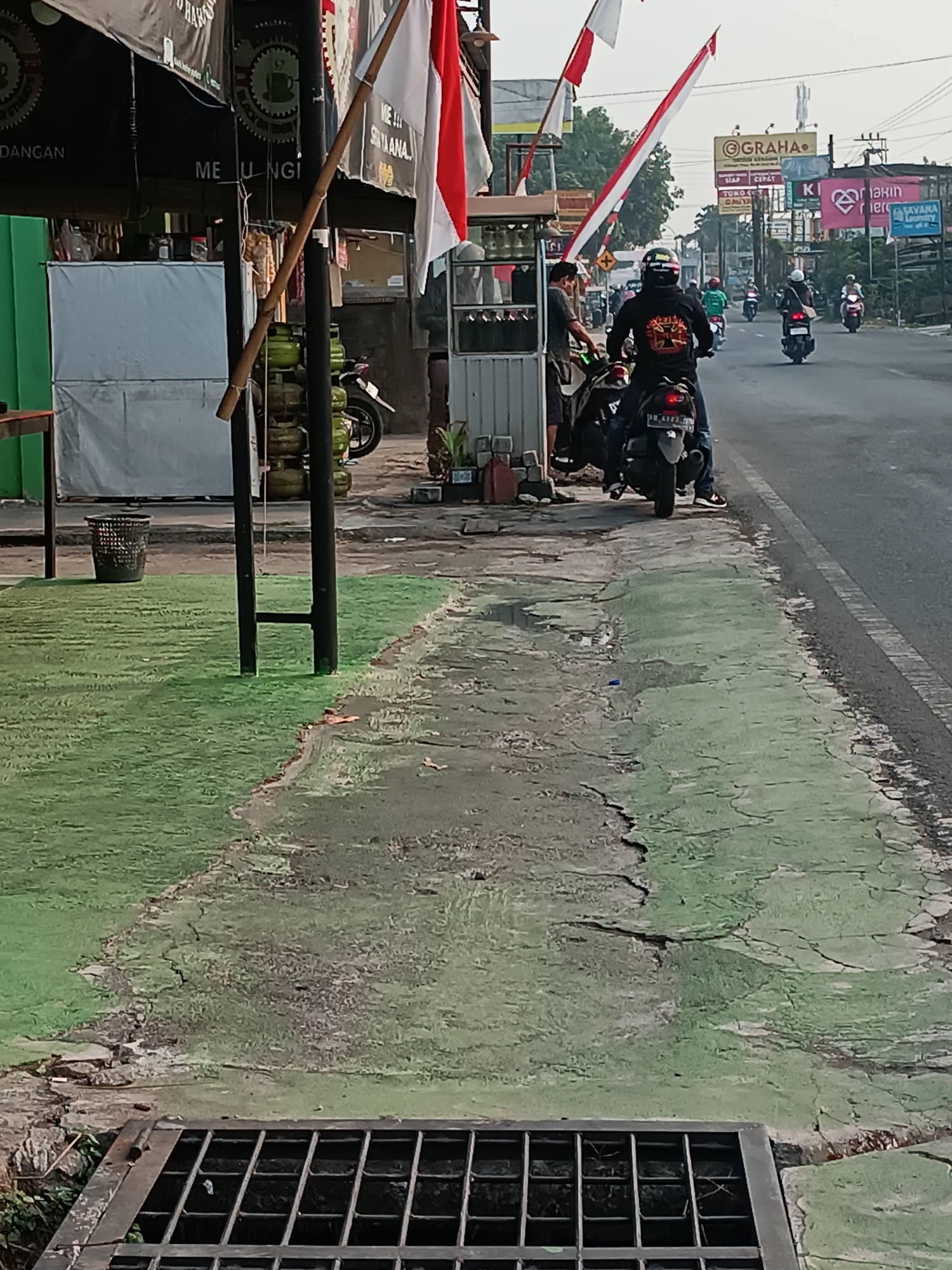 The image captures a street scene in Pleret, Indonesia, likely during daylight given the clear visibility. The foreground features a cracked, green-painted concrete surface adjacent to a metal grate over a drain. To the left, a patch of artificial green turf is visible, with a black trash bin placed on it. Further back, a black pole stands prominently, adorned with a bamboo pole and Indonesian flags. Behind the pole are stacked gas canisters.
In the middle ground, a man in a black hoodie with a graphic design on the back is seated on a motorcycle, facing away from the camera. Another person is partially visible standing near the motorcycle. The motorcycle has a license plate that reads "AB 4322 CIG". Several other motorcycles and riders are on the street, moving away from the viewer. One rider wears a helmet with colorful graphics.
The background displays a roadside environment with buildings, signage, and overhead wires. Prominent signs include "GRAHA" and "Savana Laundry". The overall impression is a bustling urban street with ongoing traffic.