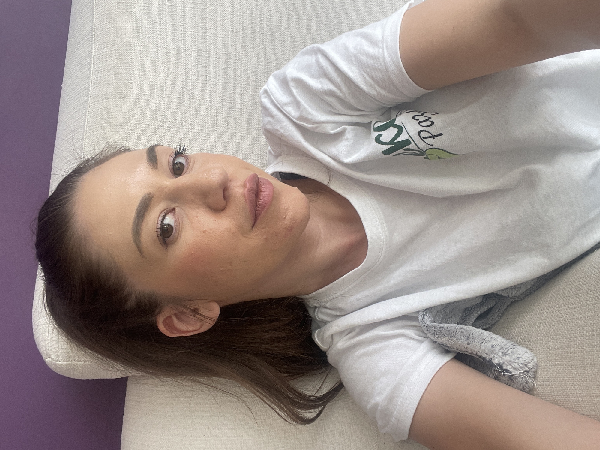A light-skinned woman with brown hair is lying on a light-colored sofa with a lilac-colored wall in the background. She is wearing a white t-shirt with green lettering, partially covered by a gray, fuzzy blanket. Her face is turned towards the camera, and she appears to be looking at the camera. There are no explicit indicators in the image that place it in Sofia, Bulgaria.