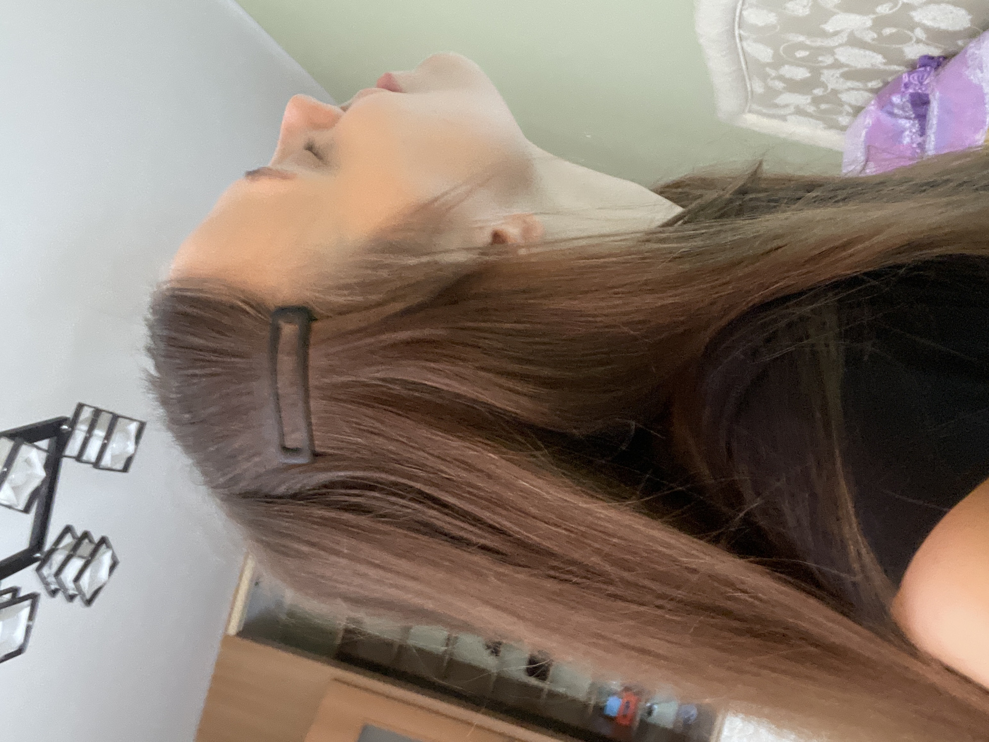 An image shows a young woman in a domestic indoor setting. The woman has long, brown hair styled with a dark brown hair clip. She is wearing a black t-shirt and is captured from the chest up, looking upwards and to the right. The background includes a wall, part of a piece of furniture with a patterned upholstery, and a ceiling light fixture. There is no visible indication of the city of Ufa, Russia, or any specific geographical location.