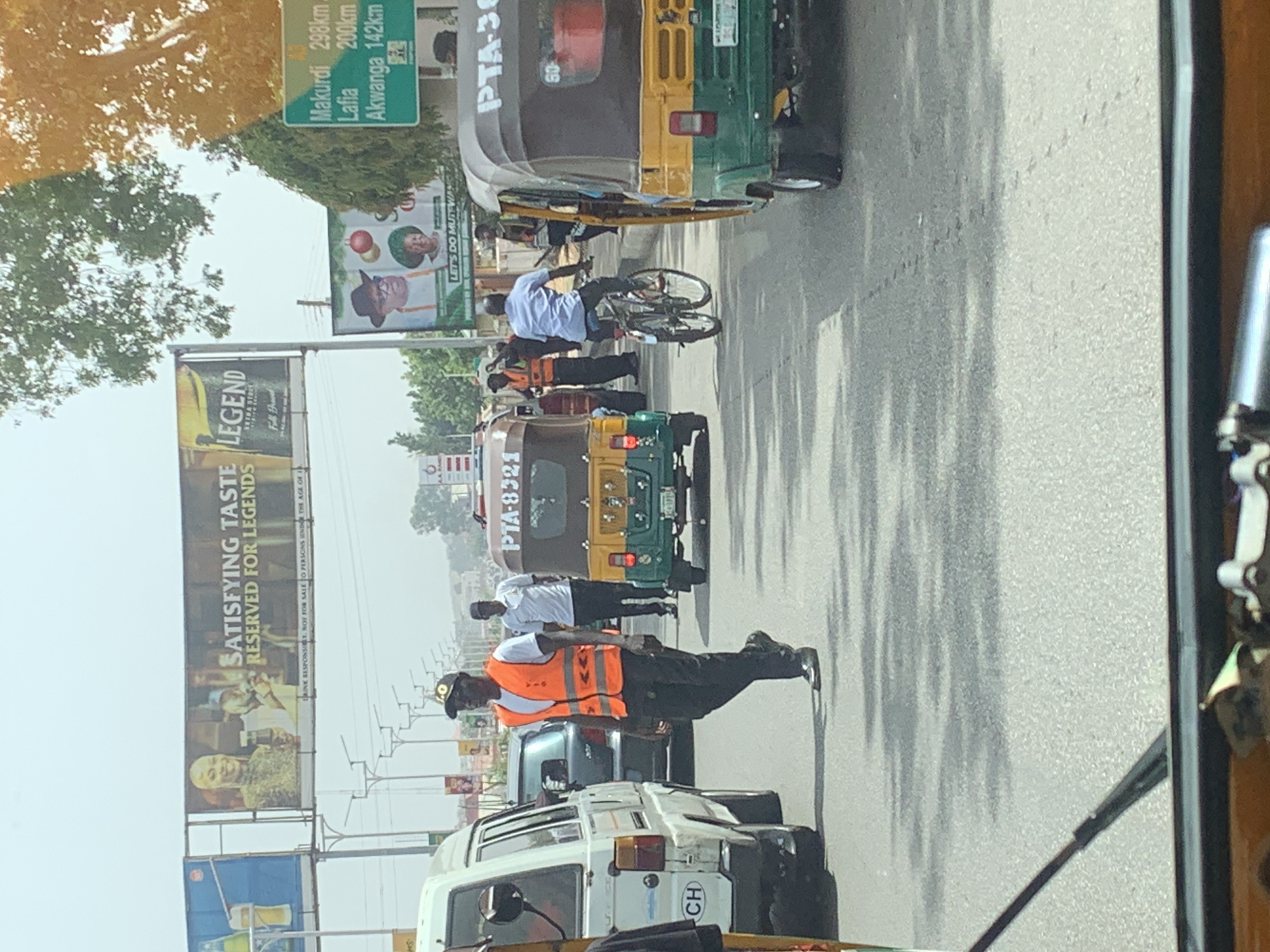 The image shows a traffic scene in Jos, Nigeria. The image is taken from inside a vehicle, looking out onto a road filled with traffic. There are commercial tricycles, a white van with the letters "CH" on it, and other cars visible. A traffic warden wearing an orange vest is standing in the road, presumably directing traffic. There is a man on a bicycle near the tricycles. Billboards are visible, one advertising Legend beer and another displaying political figures with the text "LET'S DO MUTEWA". A signpost indicates distances to various cities: Makurdi (298km), Lafia (200km), and Akwanga (142km). The weather appears to be sunny and the sky is bright.