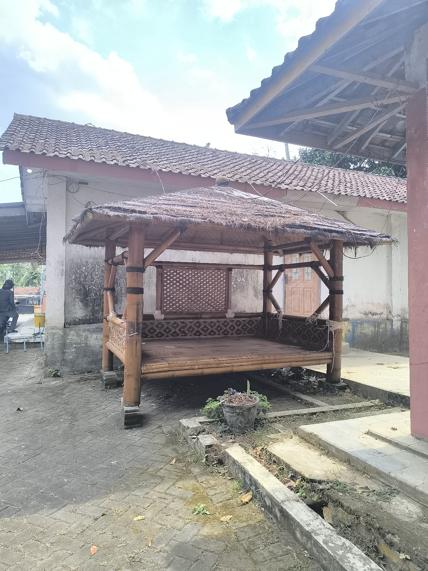 In Ranuyoso, Indonesia, a traditional bamboo gazebo with a thatched roof sits on a paved area. The gazebo features a patterned base and open sides, with bamboo poles supporting the structure. Behind the gazebo, a white building with a tiled roof is visible, along with a reddish-brown wall to the right. The ground is a mixture of brick paving and a concrete drainage channel. A person in a dark jacket and black cap is seen in the background on the left, near a yellow bucket and a blue chair. The sky is partly cloudy, with white clouds against a bright blue background.