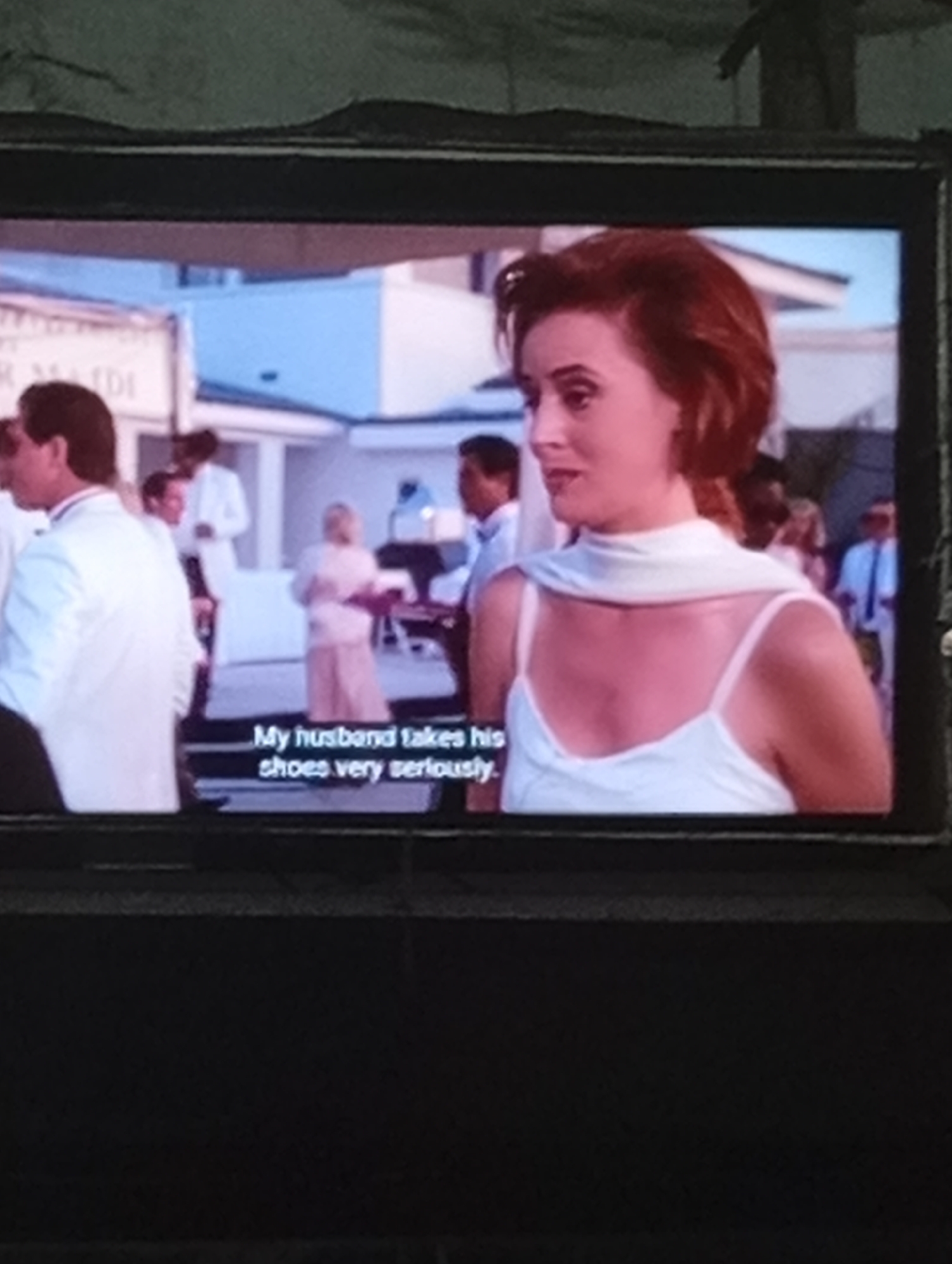 A screenshot of a television screen shows a scene from a movie or television show. In the foreground, a woman with red hair and fair skin is looking to her right and speaking. She is wearing a white camisole and a white scarf draped around her shoulders. Subtitles at the bottom of the screen read "My husband takes his shoes very seriously." In the background, several people are visible, including a man in a white suit on the left and another man in a white shirt and tie further back. A woman in a pale pink dress stands near the center. The setting appears to be an outdoor event or gathering with a building and some trees in the distance. There is no indication of the city of Lhokseumawe, Indonesia in the image.