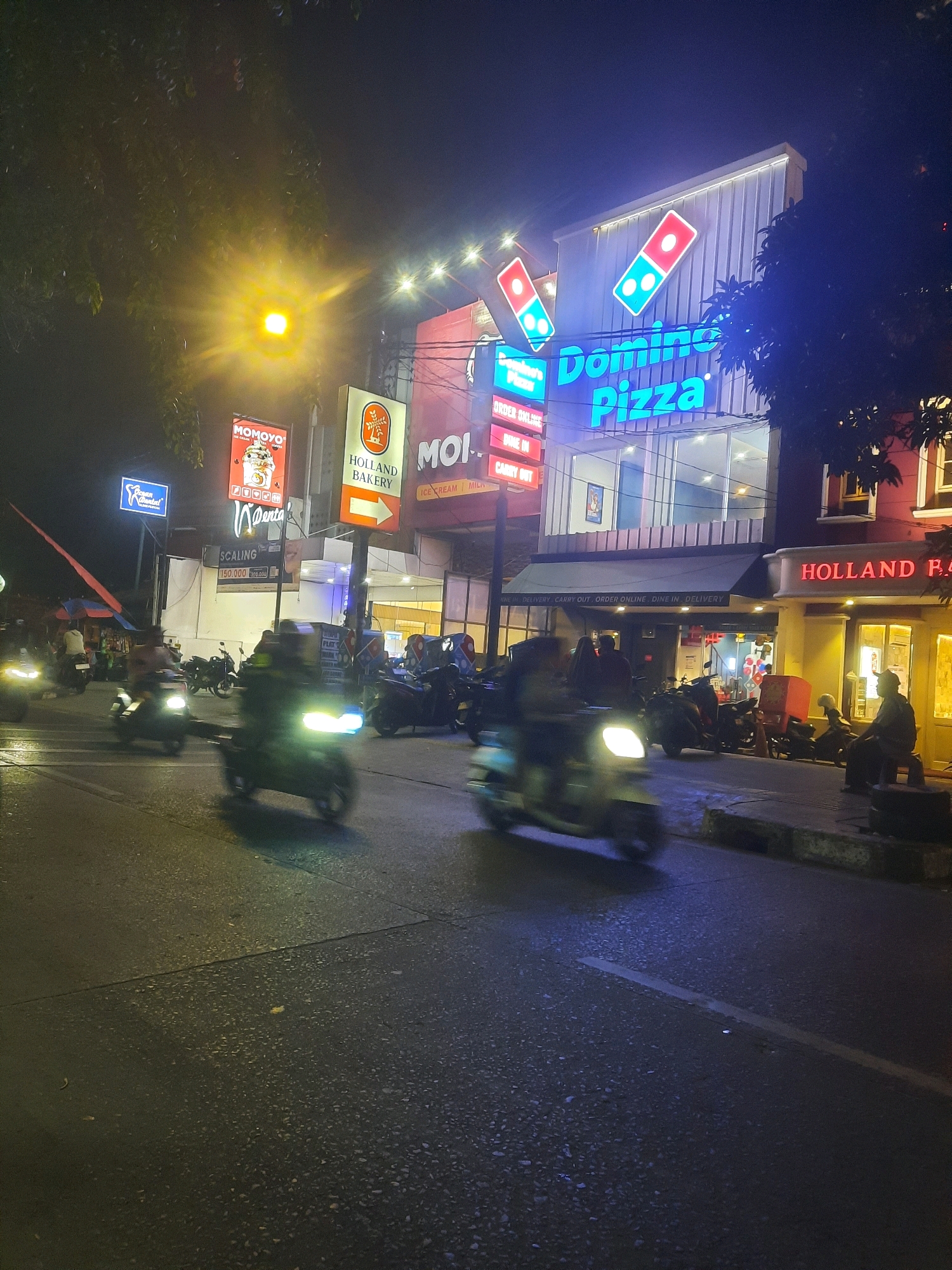 The image depicts a street scene at night in North Jakarta, Indonesia, likely on a commercial street given the numerous illuminated signs. The dominant feature is a Domino's Pizza establishment with a prominent blue neon sign. Adjacent to it, "HOLLAND BAKERY" is advertised with red lettering. Other signs visible include "MOMOYO" and "MOI ICE CREAM." The street is wet, suggesting recent rain or a humid climate, and reflects the neon lights. Several motorcycles, some with riders blurred due to motion, are present on the road, indicating active traffic. The overall atmosphere is bustling and commercial, typical of a vibrant urban setting after dark.