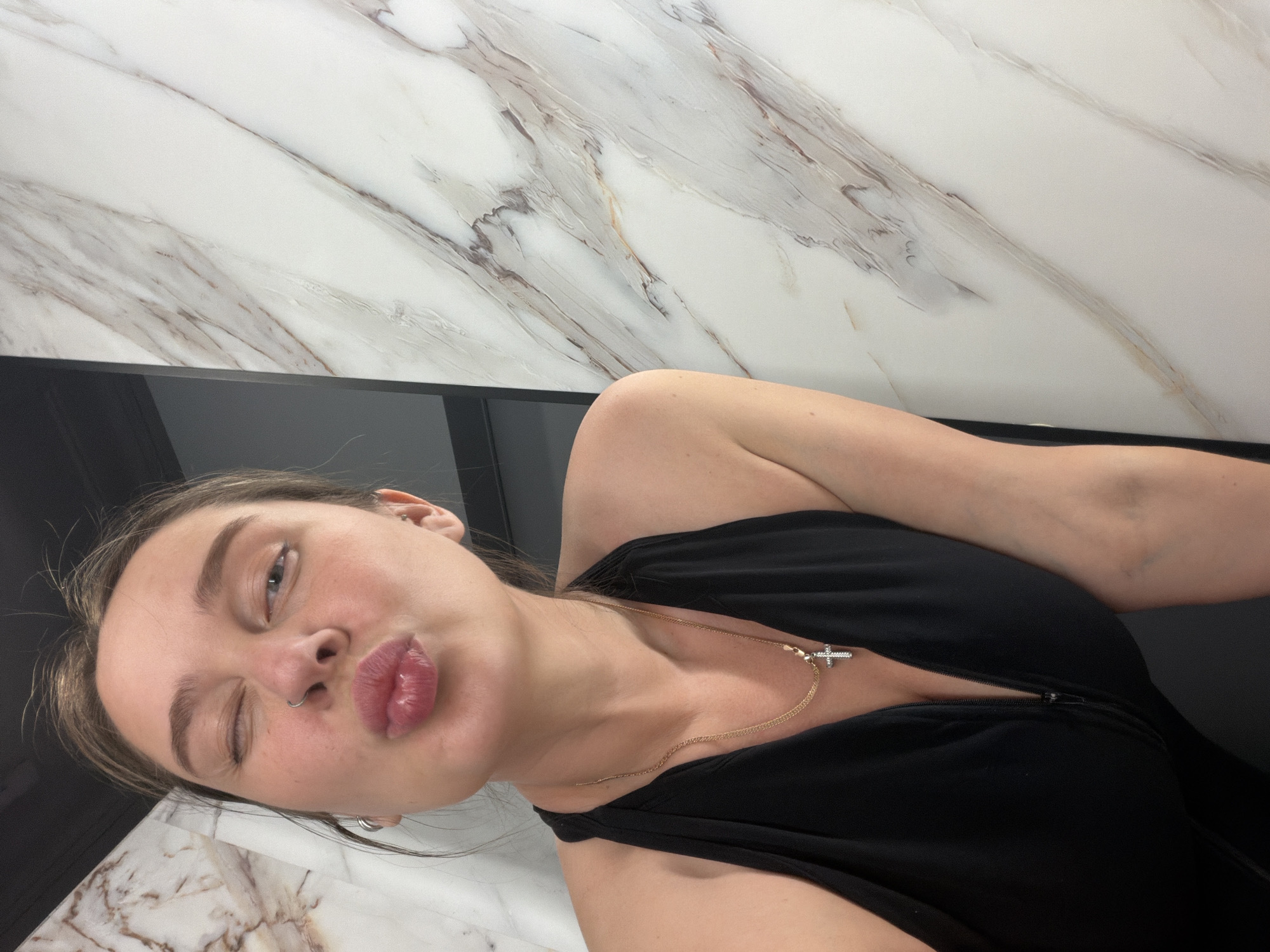 A young, light-skinned woman is captured in a selfie taken from a low angle. She has light brown hair, with some strands visible. Her right eye is closed in a wink, while her left eye is open. Her lips are pursed in a kiss. She has a nose ring and wears small hoop earrings. She is wearing a black, low-cut dress or top, revealing part of her shoulder and chest. A gold necklace with a cross pendant is visible around her neck. The background features a white marble surface with grey veining. A black surface is also visible. The image does not contain any information about the location in Moscow, Russia.
