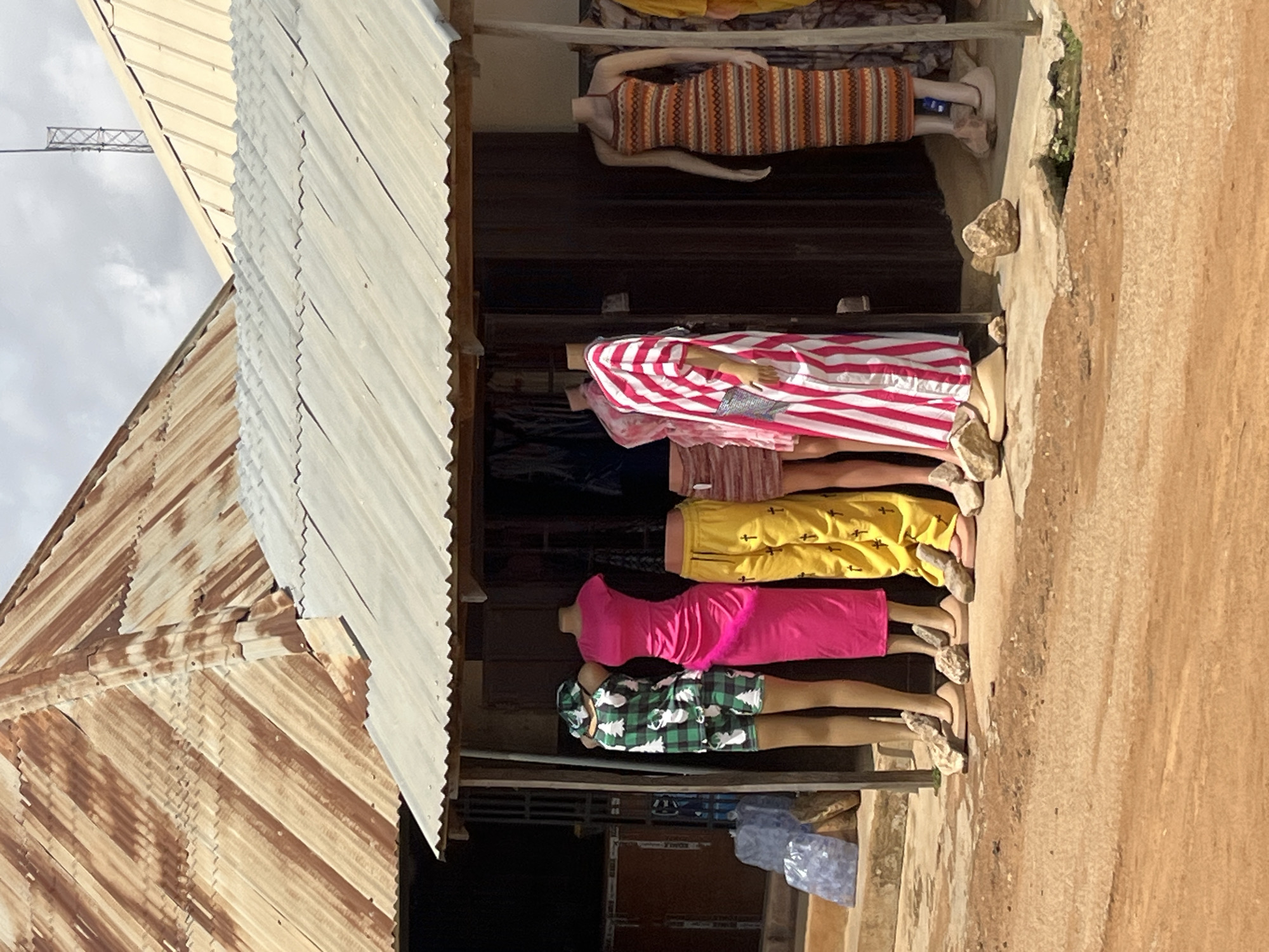 The image depicts a storefront in Bukuru, Nigeria, showcasing several mannequins dressed in various outfits. The storefront features a corrugated metal roof that appears weathered and rusty, contrasting with a lighter-colored corrugated metal awning extending over the display area. Several mannequins are positioned outside, displaying clothing items such as a green and white patterned shorts and tank top set, a vibrant pink long dress, yellow pants with a cross pattern, and a long striped dress with red and white vertical stripes. Another mannequin is visible in the background wearing a horizontally striped dress. The ground in front of the store is a dusty, reddish-brown dirt path. Scattered rocks are used as props to stabilize the mannequins' feet. In the background, several plastic bags are stacked on steps leading to a building. The sky above is overcast.