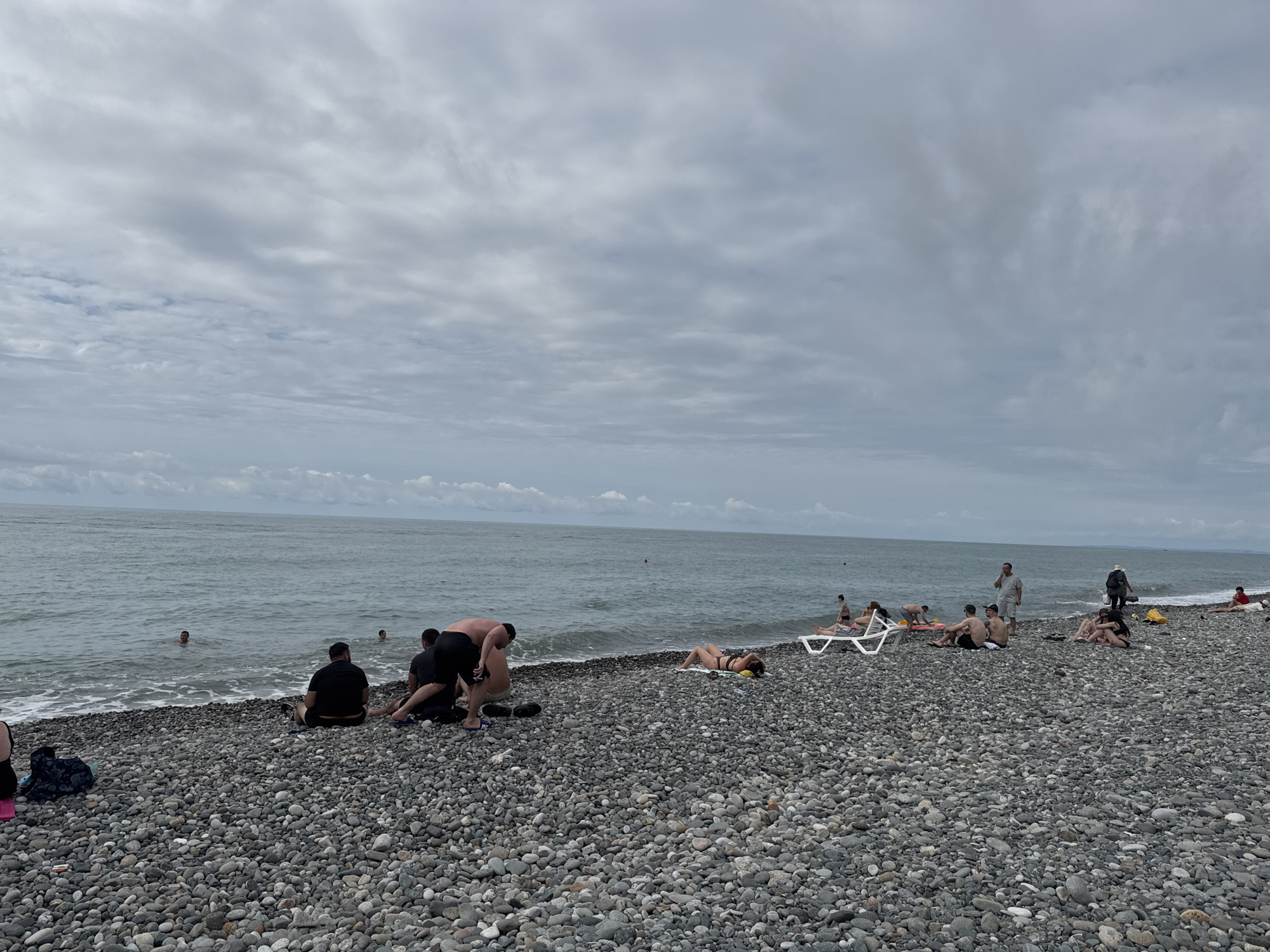The image depicts a pebble beach in Batumi, Georgia, on a cloudy day. Several people are present on the beach, some swimming in the sea, others sunbathing or sitting on the shore. The sea is calm with small waves. The beach is covered in grey pebbles. In the foreground, a few people are seated or kneeling on the pebbles. Further back, more individuals are visible, including some on lounge chairs. The horizon line is clear, separating the sea from the overcast sky.