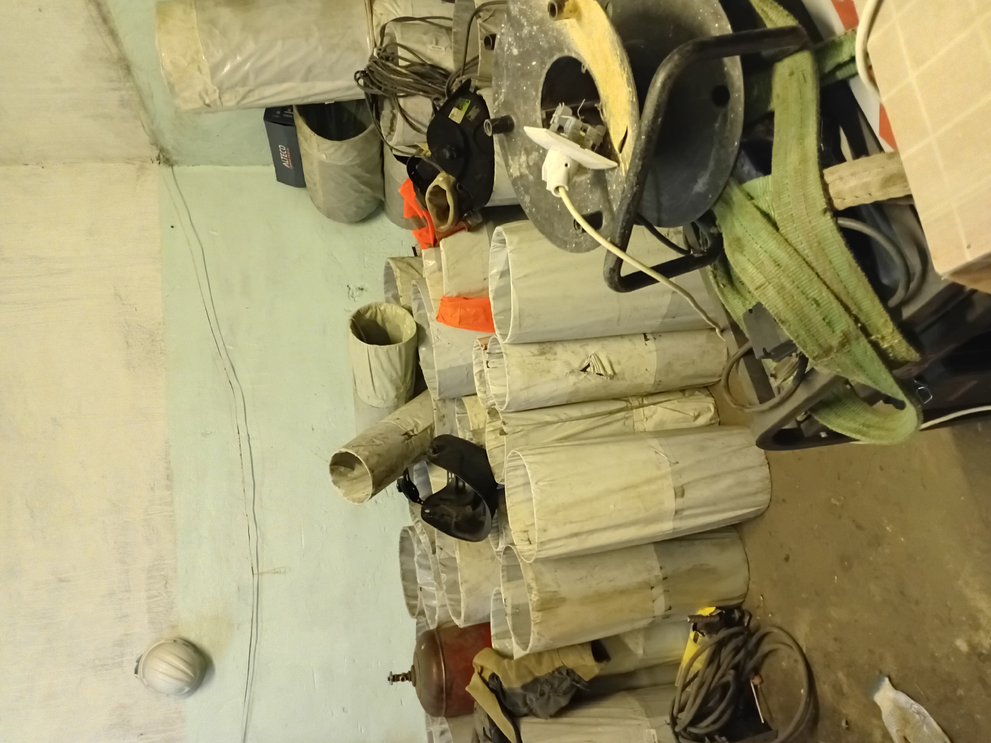 The image depicts a cluttered storage room or workshop. The walls are painted in two tones, with a lighter color at the top and a pale green at the bottom, showing signs of age and wear. Several large, cylindrical objects, possibly protective coverings or rolls of material, are stacked and leaning against each other throughout the room. Some are white and appear to be made of fabric or plastic, while others are a darker, rougher material.
In the foreground, a black reel with a yellow rim is prominently displayed, with a white electrical cord plugged into it. A green strap is draped over some equipment behind the reel. To the left, a coiled black cable lies on the floor, alongside what appears to be a work glove.
Further back, a red propane tank with a brass valve is visible. A black welding mask is placed on top of some of the cylindrical objects. Bright orange safety cones or vests are also scattered among the items. A white hard hat is affixed to the upper wall on the left side of the frame. In the background, a dark blue and grey box with the label "ALTECO" is visible. The floor is concrete and appears dusty. The overall impression is one of a functional but unorganized space, likely used for storage or work related to industrial or construction activities. The location is identified as a storage room in Semey, Kazakhstan.