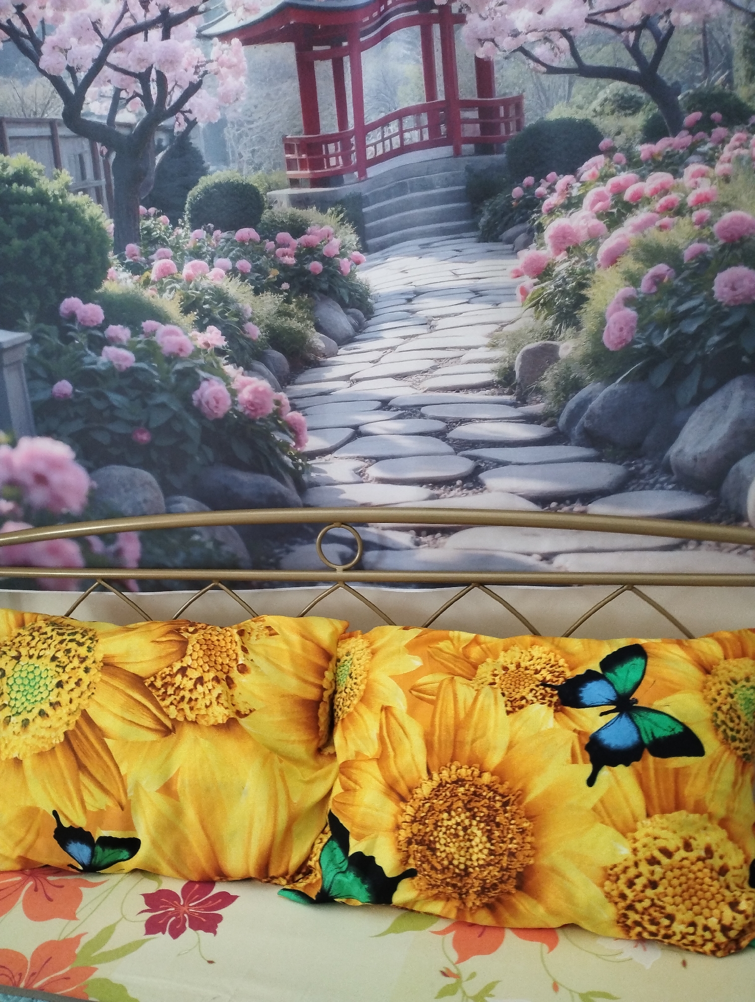 The image shows a close-up of a bed with two pillows and a headboard against a wallpaper. The wallpaper depicts a picturesque Japanese garden scene with a winding stone path leading to a red pagoda-style building. Cherry blossoms in full bloom adorn trees on either side of the path, and pink flowers line the pathway. The pillows are covered in a vibrant yellow fabric featuring large sunflower prints and small blue and green butterflies. The headboard is a gold-colored metal frame. The overall impression is one of a serene and floral aesthetic.