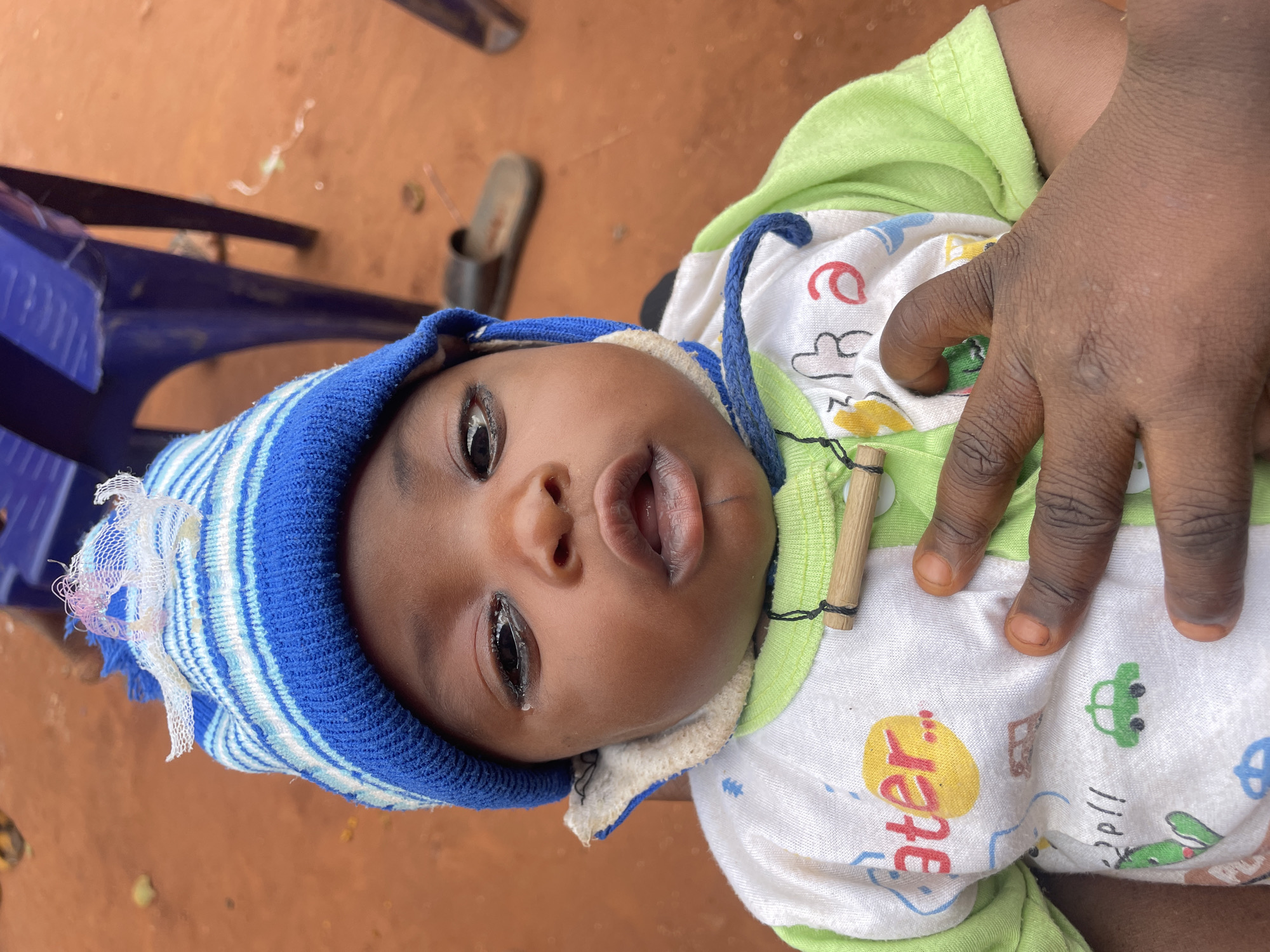 A baby lies on their back, wearing a blue and white striped hat and a white onesie with green trim, featuring cartoon images and the word "later." The baby's face is centered in the frame, with their eyes open. A dark-skinned adult hand is partially visible over the baby's chest. The background is reddish-brown, possibly dirt or a floor, with the leg of a blue plastic chair and a sandal visible in the upper left. The location is Ogbe, Nigeria.