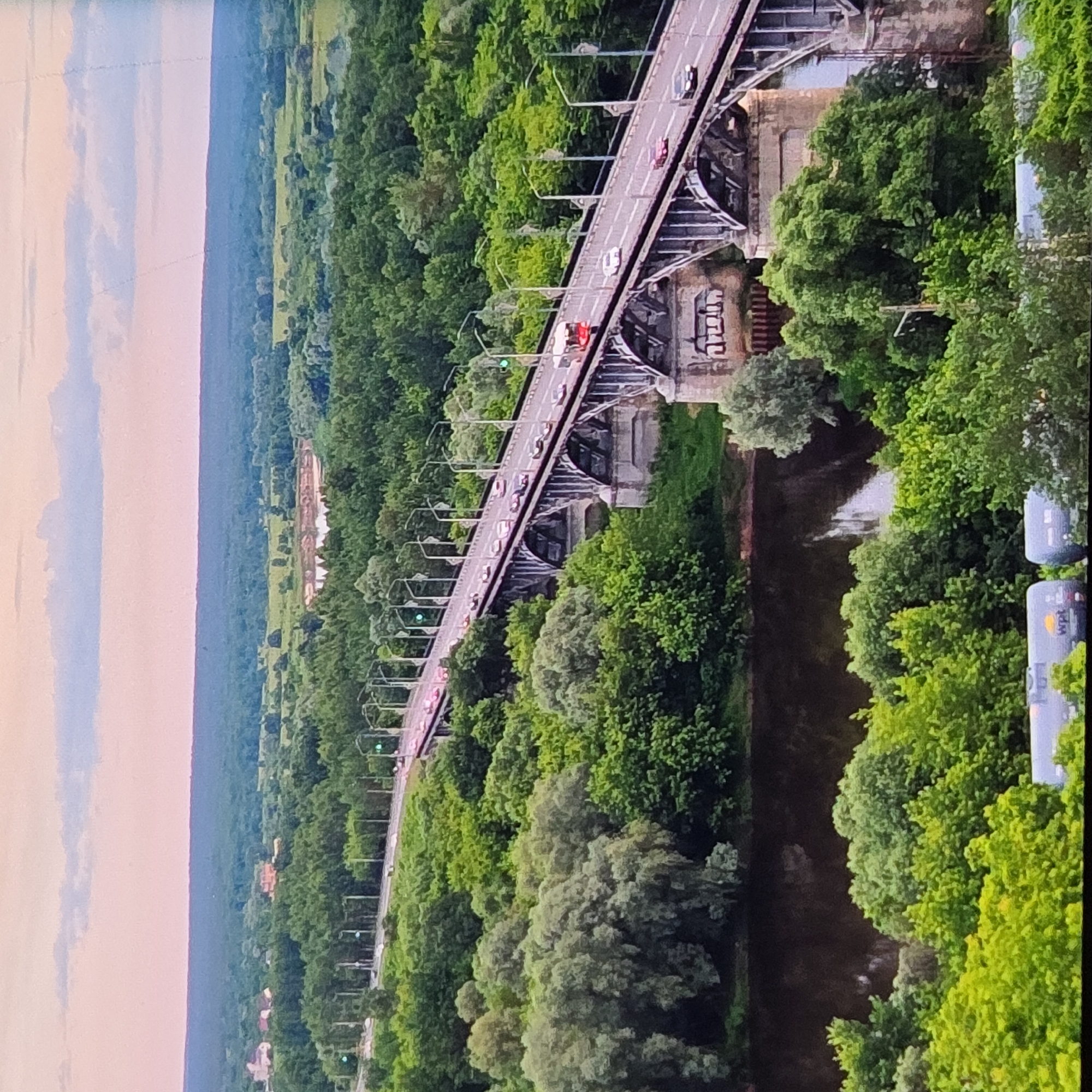 This image depicts a wide-angle, aerial view of the bridge over the Klyazma River in the city of Vladimir, Russia. The bridge, a prominent concrete and metal structure with multiple arches, carries a two-lane road with a moderate amount of traffic, including cars and a red bus. The road is lined with streetlights, some of which are illuminated with green signals. The bridge is surrounded by lush, dense green trees on both banks of the river. The Klyazma River itself is visible beneath the bridge, appearing dark and calm. In the background, the landscape consists of rolling hills covered in more trees, with a small lake or pond visible in the distance. The sky is a soft pink and blue gradient, suggesting either sunrise or sunset. In the foreground, at the bottom of the image, two gray train tank cars are partially visible, one with the word "wpt" printed on it. The overall scene is one of urban infrastructure integrated with a natural, verdant environment.