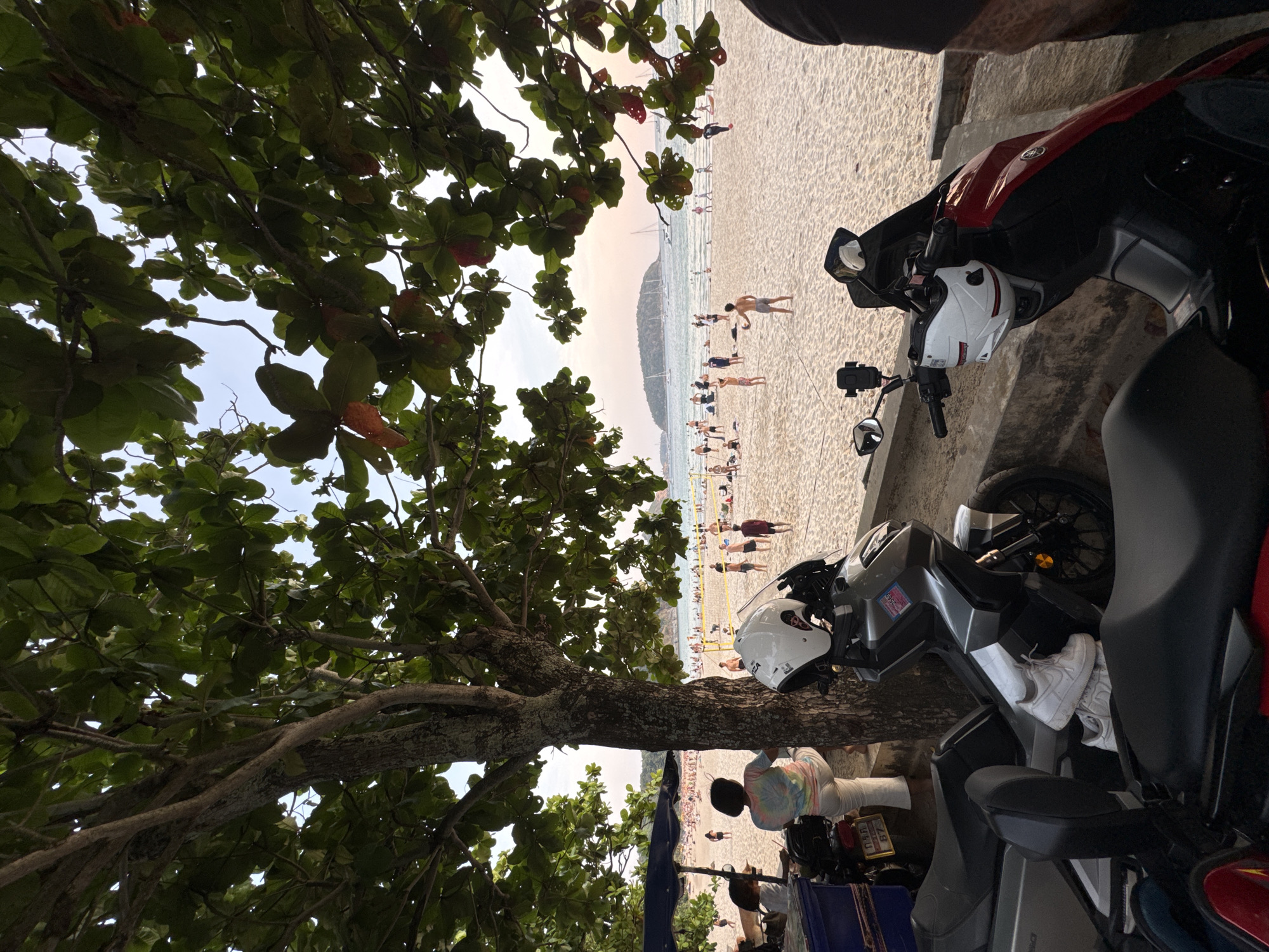 The image shows a beach scene in Rawai, Thailand. A tree with green leaves frames the left side of the image. In the center background, there is a sandy beach with people engaging in various activities such as sunbathing, swimming, and playing volleyball. In the foreground, there are two parked scooters with helmets placed on them, indicating a mode of transportation for accessing the beach. A person wearing white sneakers is seen standing next to one of the scooters. A vendor or stall is partially visible in the lower portion of the frame, and a person appears to be working in it. The overall environment suggests a popular beach area with a mix of locals and tourists enjoying recreational activities.