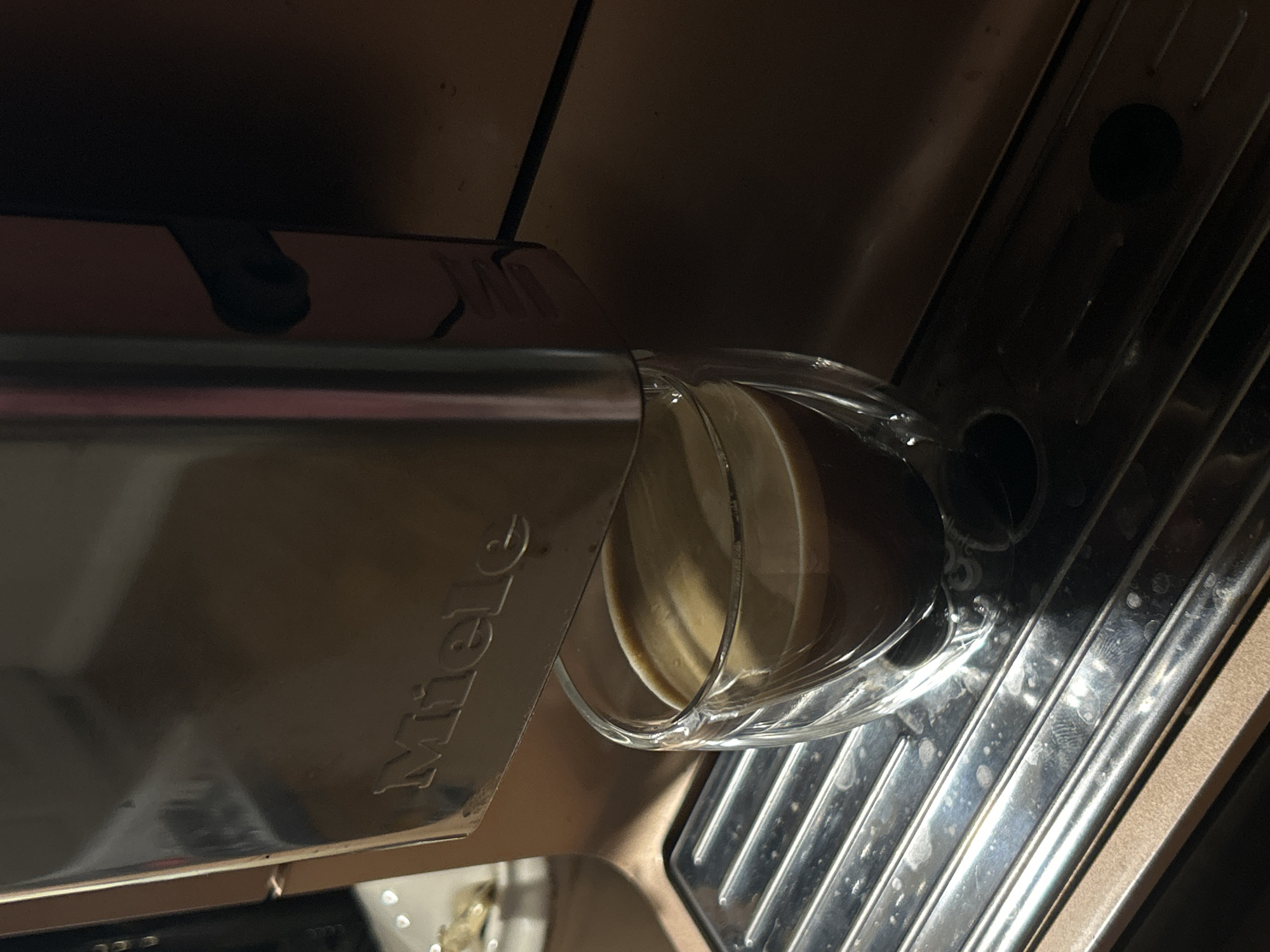 An off-kilter close-up shot depicts a Miele coffee machine dispensing espresso into a clear glass cup. The machine is chrome. A drip tray of metal grill is directly below the cup. The background is blurry and out of focus. The location and city are not present in the image.