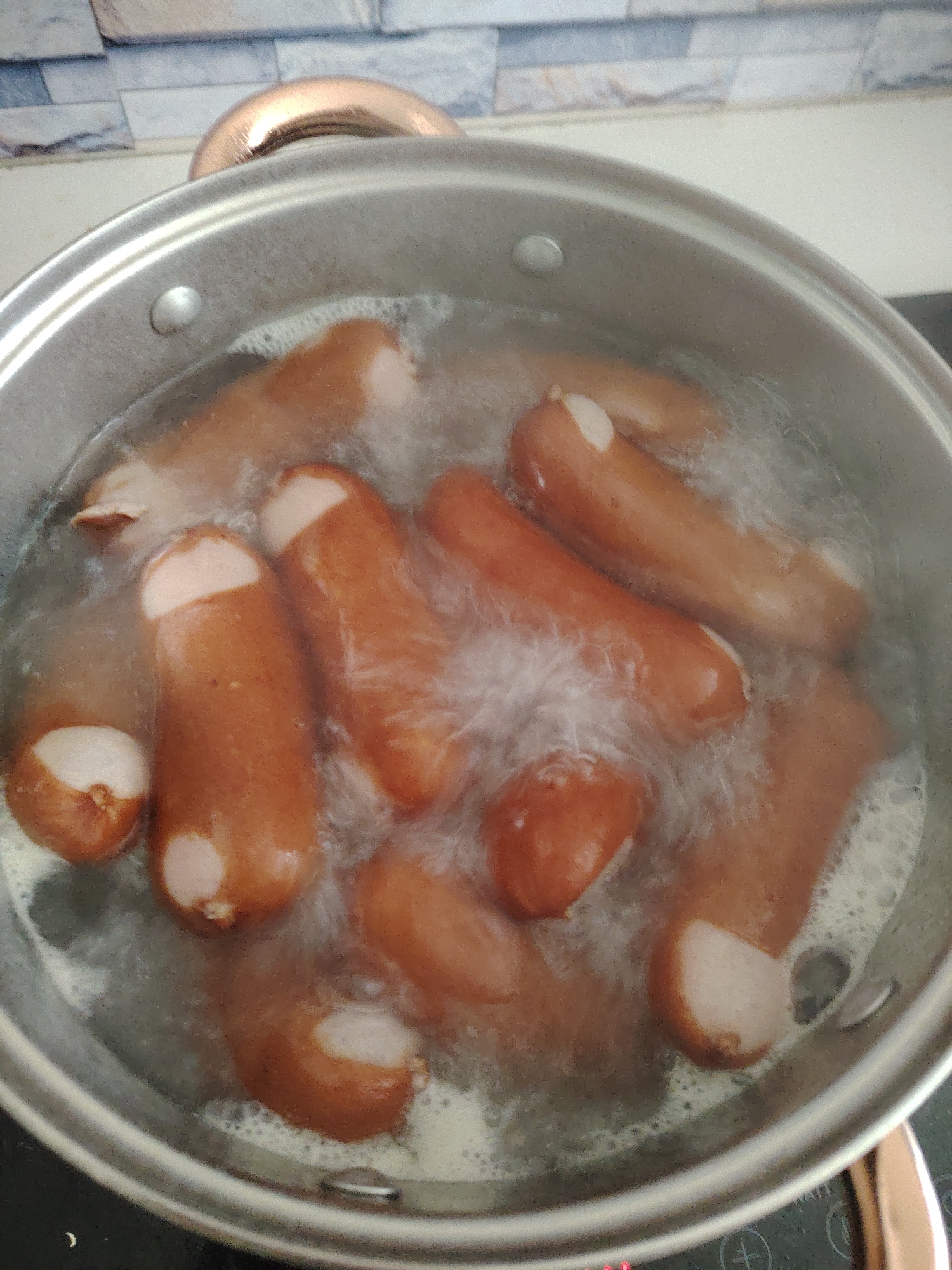 The image displays an overhead view of a silver-colored metallic cooking pot containing approximately ten to twelve frankfurters or sausages actively boiling in water. The water is effervescent with numerous bubbles and visible steam. Many of the light brown sausages show pale, expanded ends where their casings have split due to cooking. A single rivet is visible on the upper interior wall of the pot. In the upper background, part of a copper-toned pot handle is visible, likely from the lid or another part of the pot. The background features a tiled kitchen wall composed of irregularly shaped, rectangular tiles in various shades of grey, white, and light brown. To the lower right, a dark, flat stovetop surface is visible, showing faint white control panel markings and a red illuminated indicator light. The scene depicts food preparation within an indoor kitchen environment. There is no visual information within the image to determine its specific geographical location, such as the city of Huntingdonshire, United Kingdom, or any other region.