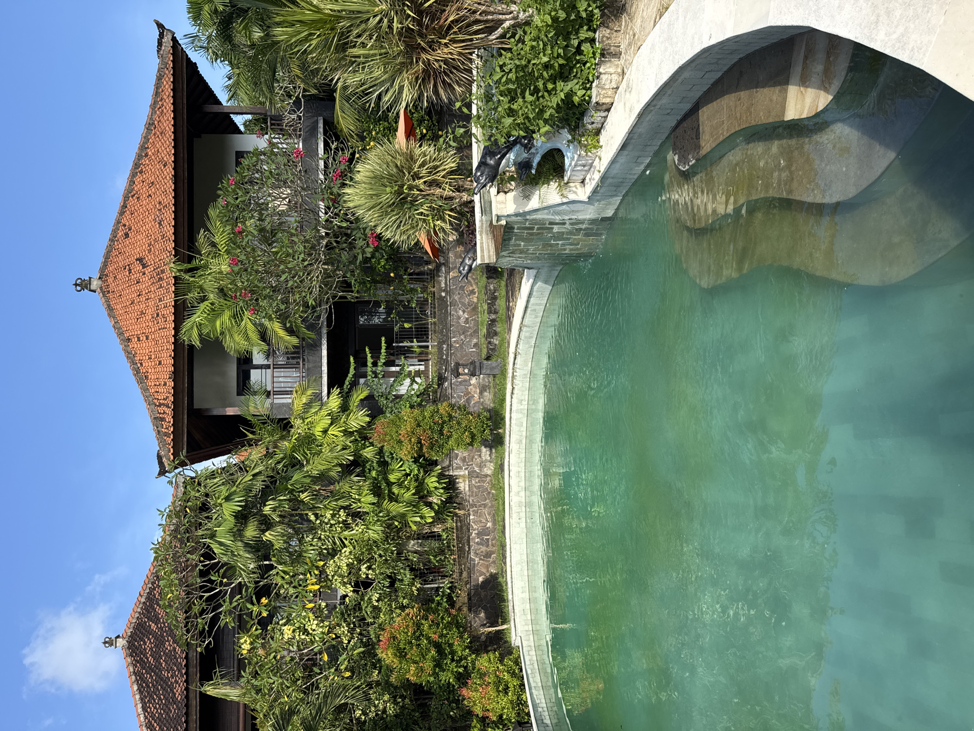 The image depicts a view of a pool and surrounding landscape in Ungasan, Indonesia. The pool is curved with multiple levels of steps leading into the water, which has a green hue. The pool is surrounded by lush greenery, including trees, bushes, and flowering plants. A building with a red tile roof is visible in the background, partially obscured by the foliage. A stone pathway or wall borders the vegetation near the pool.