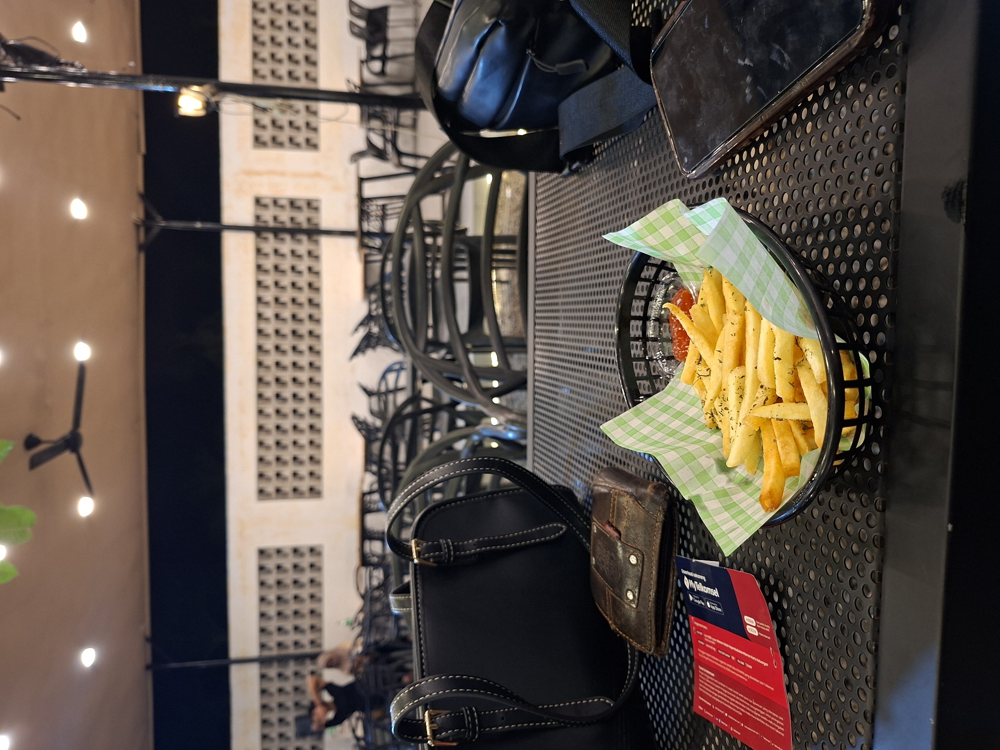 The image captures an evening scene at an outdoor or semi-outdoor dining area, likely a cafe or restaurant, in Lhokseumawe, Indonesia. The primary subject is a perforated black metal table, presented sideways due to the photo's orientation. On the table, a black basket holds a serving of golden-brown french fries seasoned with herbs, accompanied by a small container of ketchup, all resting on a green checkered paper liner.
Beside the fries, a dark-colored smartphone lies on the table, along with a black small bag or backpack and a worn brown leather wallet. A red card is also visible on the table.
In the blurred background, several buildings with distinctive square grille patterns are visible. The setting is illuminated by numerous string lights, a ceiling fan with lights, and streetlights, indicating it is nighttime. Faint, out-of-focus figures of people and empty chairs suggest other patrons or seating arrangements in the distance.
**Visible text:** The red card clearly displays "MyTelkomsel," "Download sekarang" (Download now), and the logos for Google Play and App Store. Smaller text in Indonesian includes phrases such as "Dapatkan internet seru" (Get exciting internet), "beli pulsa" (buy credit), and "bayar tagihan" (pay bills), indicating services from the telecommunications company Telkomsel.