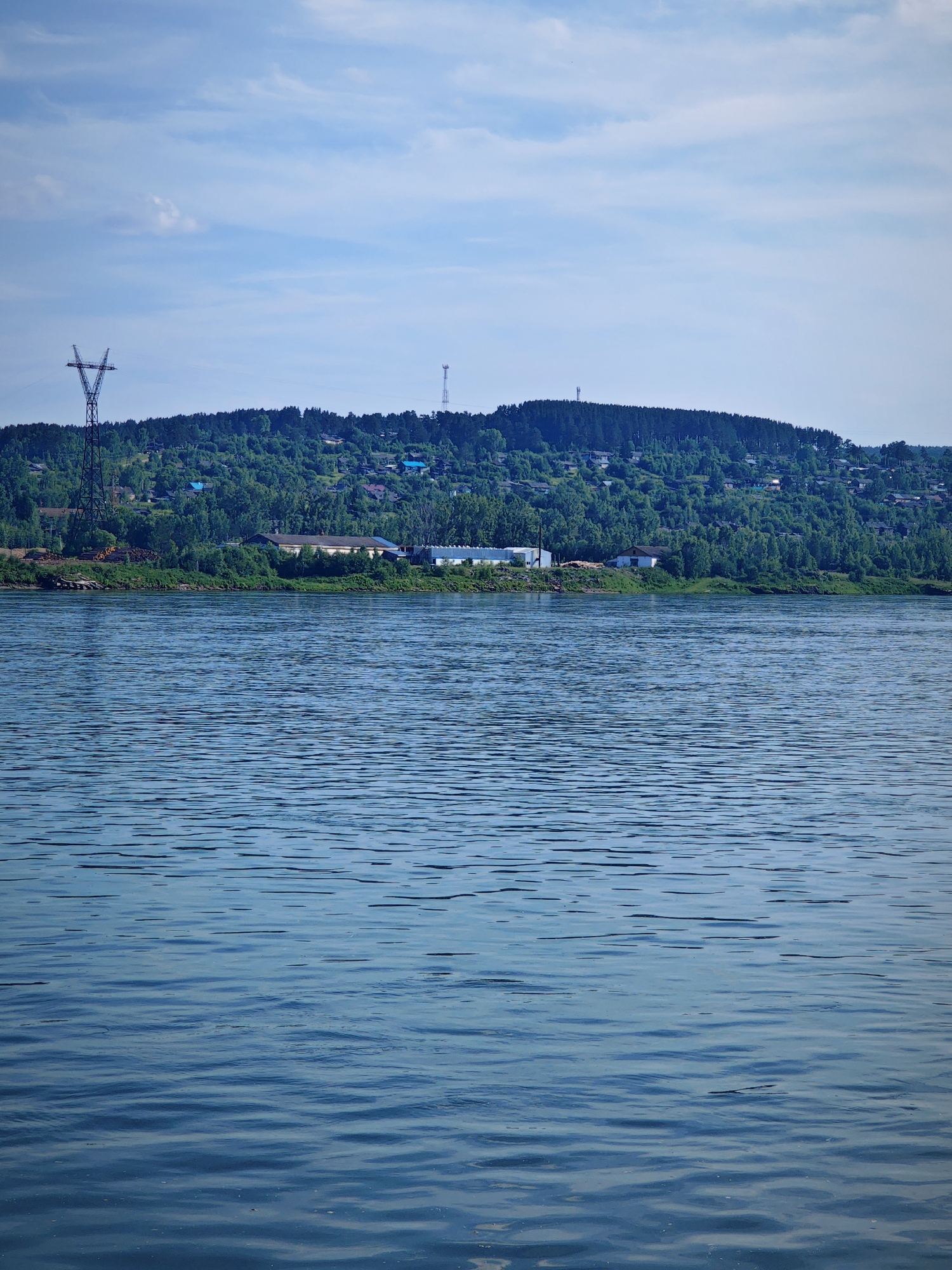 This is a vertical, eye-level, full shot of a wide, blue body of water with a shoreline and a wooded hill in the background. The water is rippled and reflects the cloudy sky. Along the shoreline, there are some buildings, including what appears to be an industrial facility and several houses with blue roofs. A tall, metal power line tower stands on the left side of the frame, and a cell tower is visible on the top of the hill. The hill is covered in dense green trees, with more houses scattered among them. The sky is blue with scattered white clouds. This scene is characteristic of the city of Predivinsk, Russia, situated on the Yenisei River.