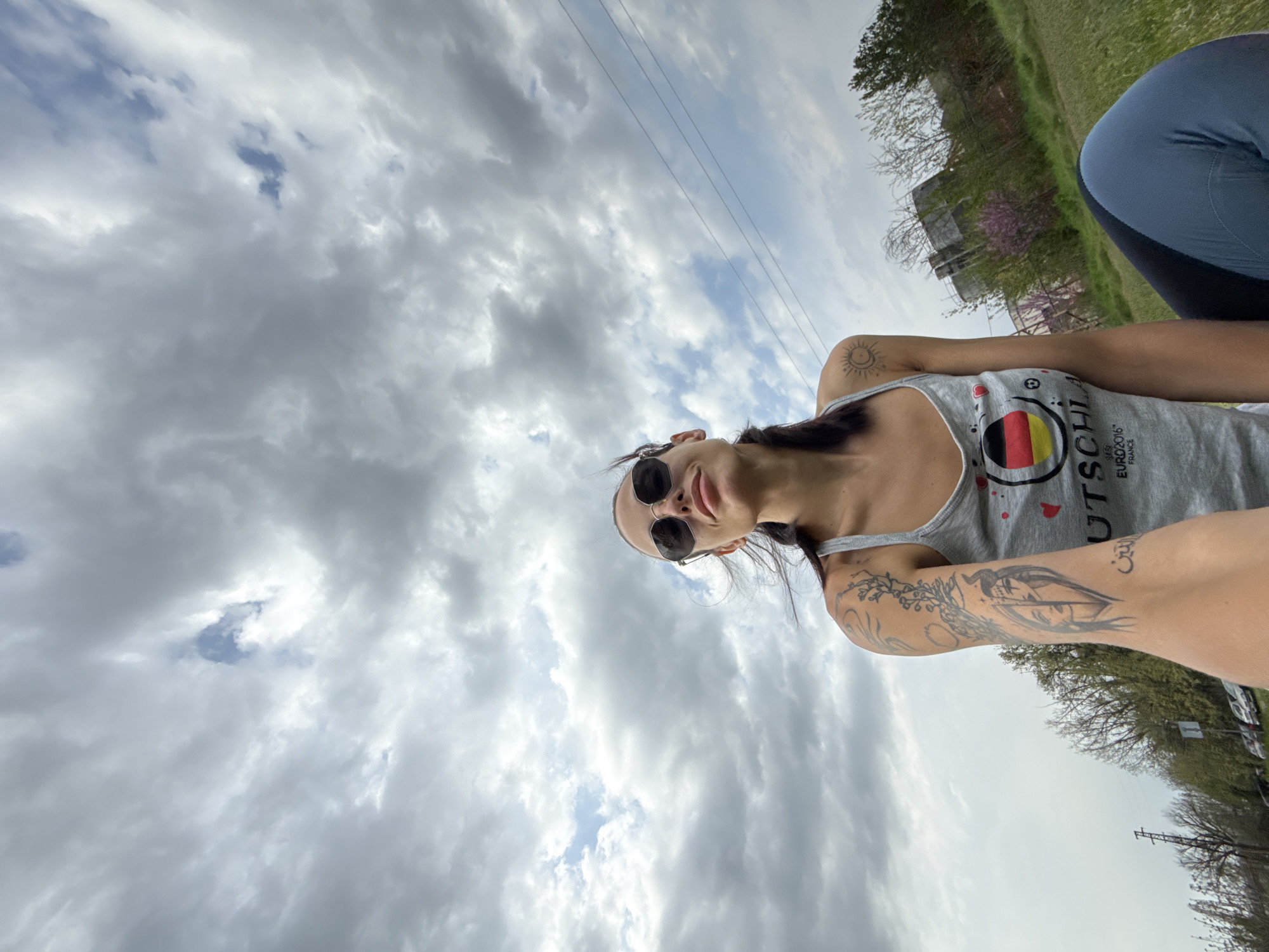 A low-angle outdoor selfie captures a person looking up towards the camera against a dramatic, cloudy sky in Pleven, Bulgaria. The sky is predominantly filled with heavy grey and white clouds, with intermittent patches of blue visible. Overhead power lines traverse the frame.
The main subject, a female, is positioned in the foreground, with her head and upper torso visible. She has dark hair, wears sunglasses, and looks downwards towards the camera with a subtle, relaxed smile. She is wearing a grey tank top featuring the word "DEUTSCHLA" alongside a German flag design (black, red, yellow), with "EURO2016" and "FRANCE" also visible below it, indicating a Germany Euro 2016 football shirt.
Her arms are adorned with tattoos: a stylized sun on her left shoulder/upper arm and intricate designs, including what appears to be a face or eyes, on her right forearm, along with script that resembles Arabic or Persian.
To the far right, a second person or part of a person is partially visible, wearing blue and black athletic attire. The background consists of trees with sparse foliage, suggesting early spring or late autumn. Further back, industrial-like structures, possibly water towers, are partially obscured by the trees. The overall impression is a casual outdoor moment on an overcast day.