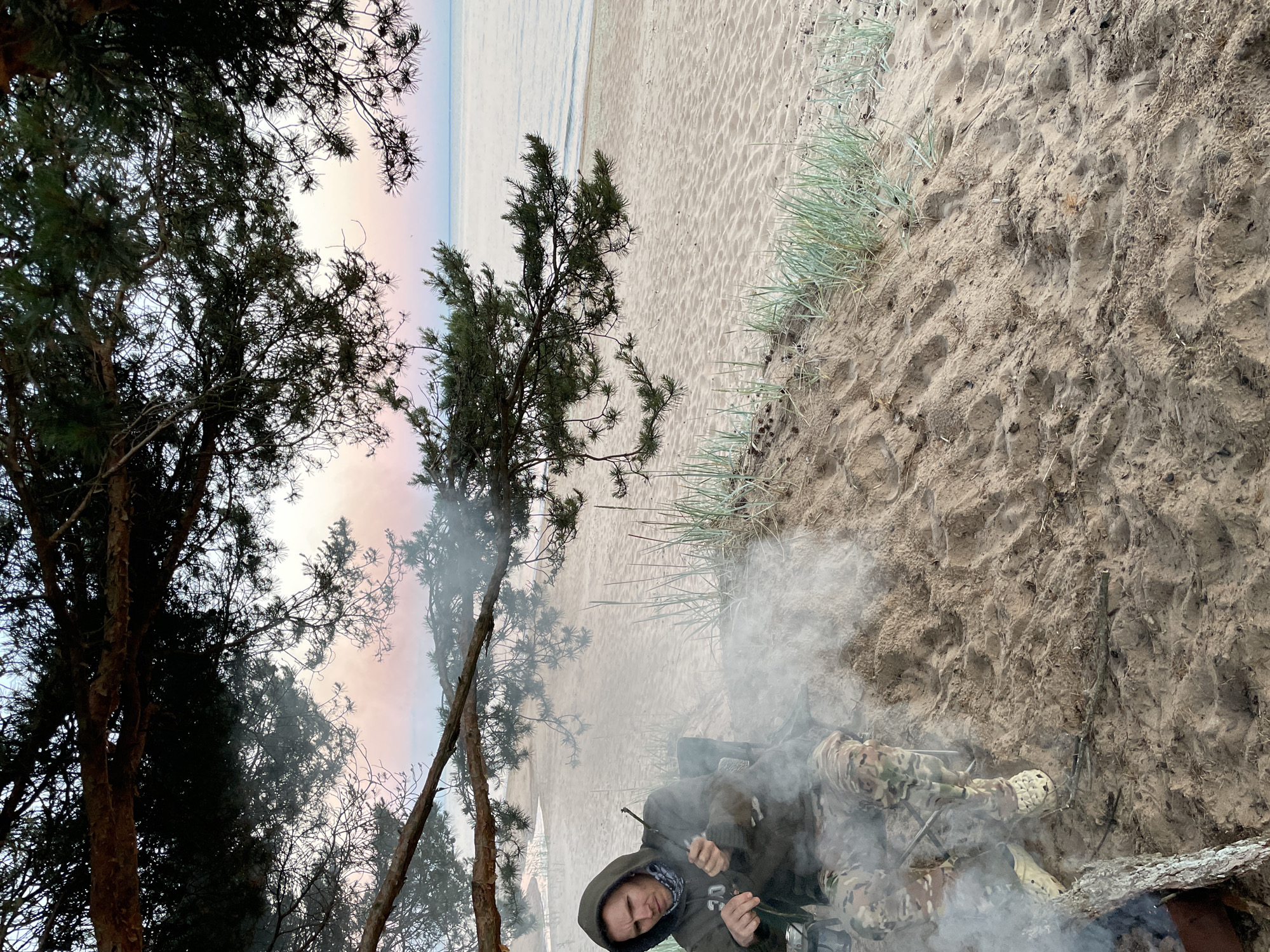 A lone Caucasian male is seated on a camping chair on a sandy beach, facing left and holding a stick over a small campfire that is producing a significant amount of smoke. He is wearing a dark hooded sweatshirt with camouflage pants and light-colored clogs. The background features a calm body of water under a sky with pink and blue hues, likely indicating dusk or dawn. Pine trees with dark green needles overhang the scene from the top and left. Grass and sand dunes are visible on the beach. The man appears to be tending to the fire or preparing something with the stick. The overall environment suggests an outdoor recreational setting.