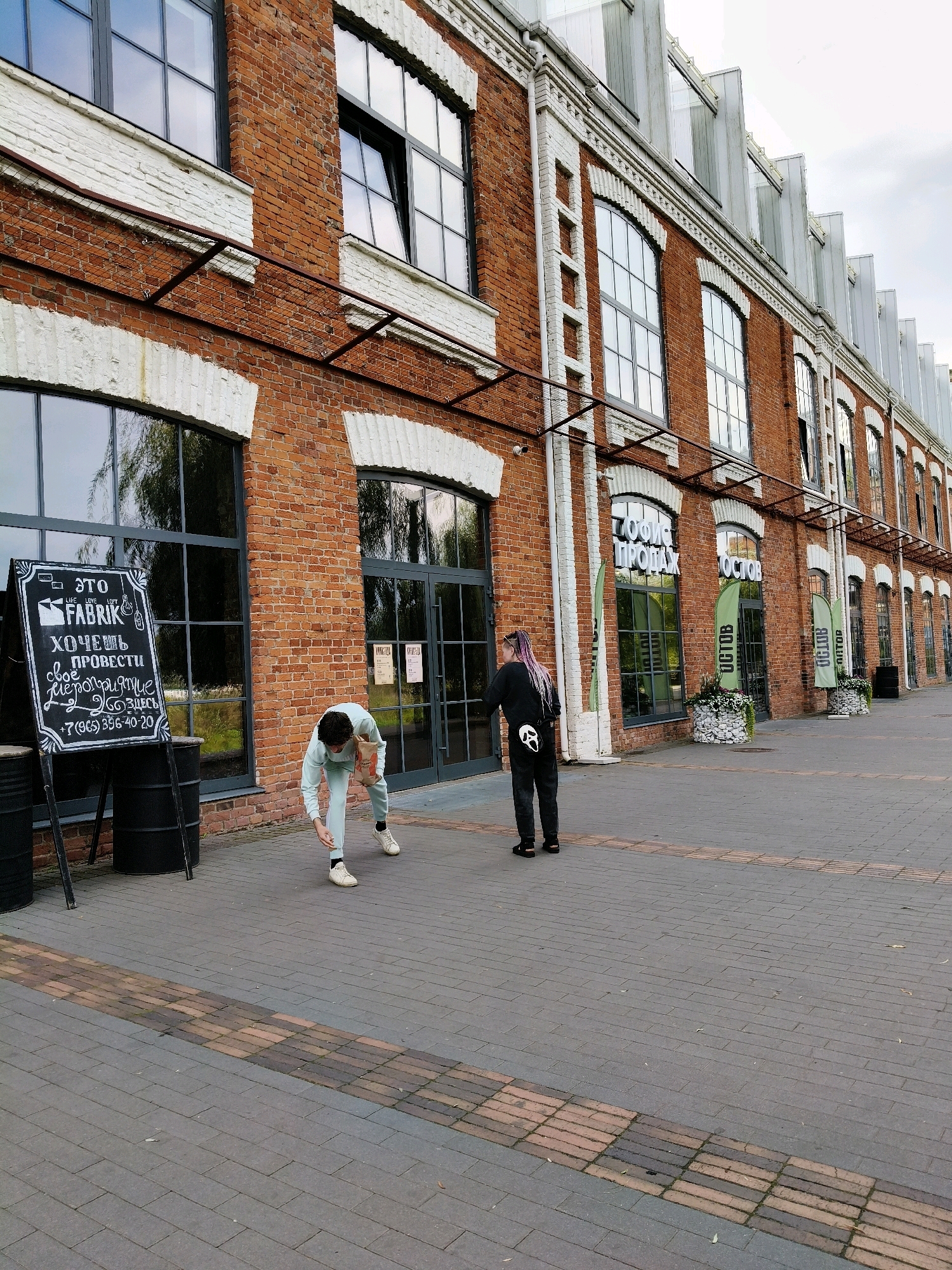 The image captures the exterior of the "Fabrik" complex, located in Naro-Fominsk, Russia. The building is characterized by its red brick facade and large, industrial-style windows. In the foreground, two individuals are visible on a paved walkway. One person, wearing a light blue tracksuit, is bent over, seemingly picking something up from the ground. The second individual, with long purple hair and dressed in black, stands facing away from the camera, looking towards an entrance. A blackboard sign with text in Russian, possibly advertising or providing information about "Fabrik", stands to the left of the frame. The walkway is made of brick pavers, with a distinct pattern of alternating colors. The environment suggests an urban setting, possibly a cultural or commercial space.