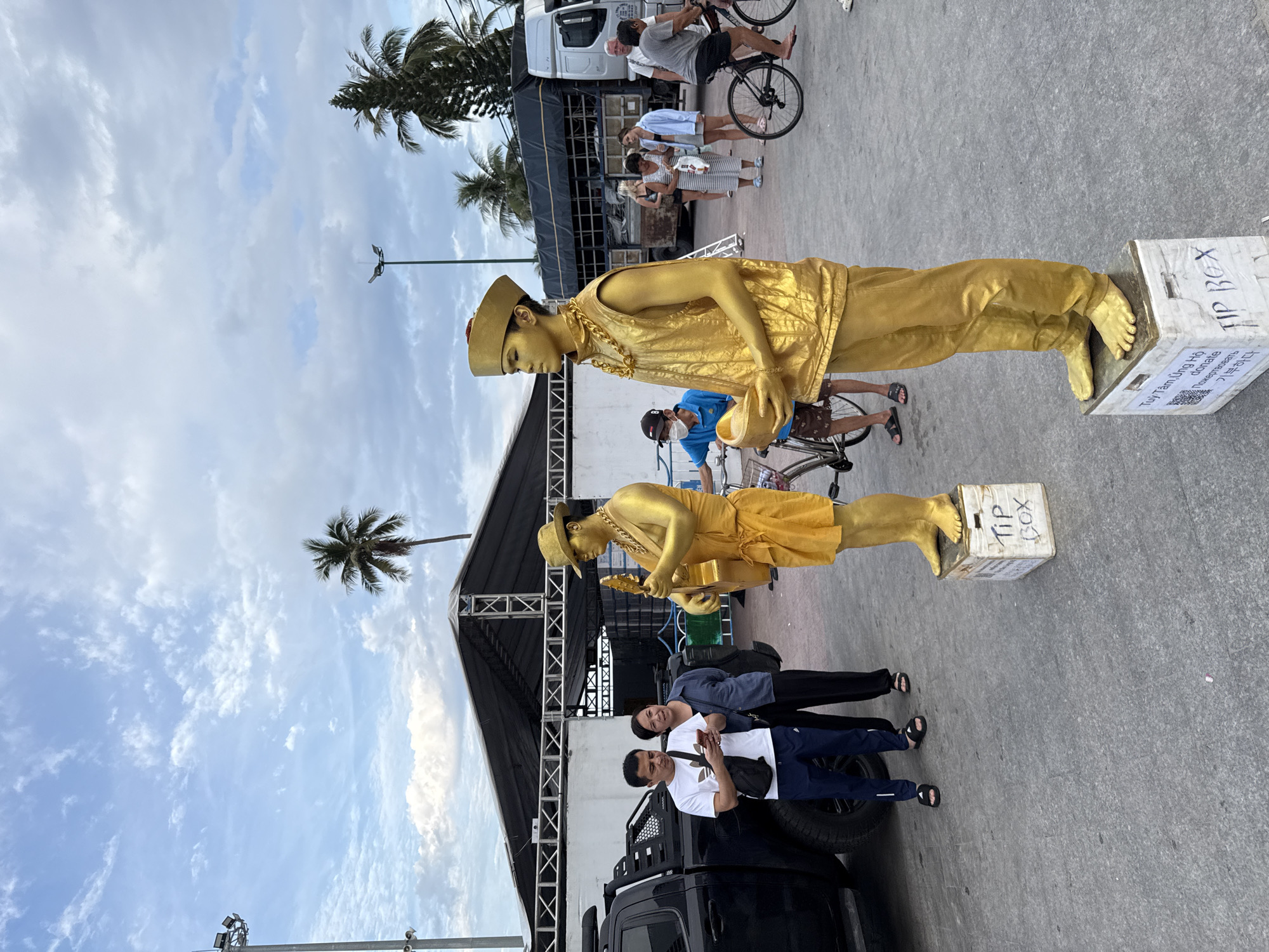 The image depicts a street scene in Nha Trang, Vietnam, with two performers painted gold standing on white boxes. One performer, dressed in a gold vest and hat, holds a guitar, while the other, wearing a gold cap, vest, and pants, holds a gold bowl. A couple is looking at their phones near a black truck. In the background, several people are walking and cycling, and a large truck is parked. The sky is partly cloudy with palm trees visible. The scene appears to be set up for public entertainment or donation collection, as indicated by the "TIP BOX" labels on the performers' boxes.