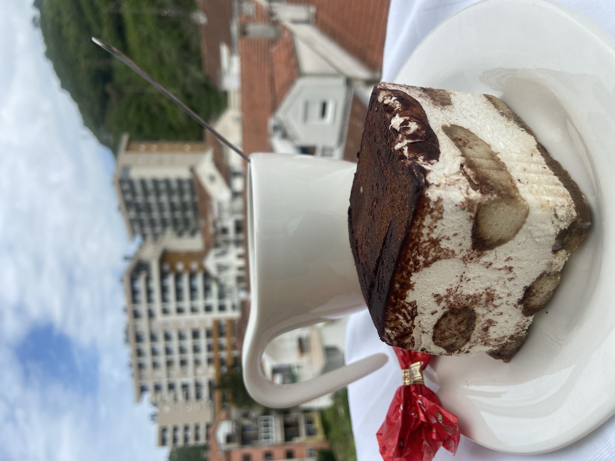 A close-up, tilted, outdoor shot shows a white coffee mug, a white plate with a slice of tiramisu, and a red-wrapped candy. The background reveals a cityscape of Budva, Montenegro. The mug has a spoon in it. The tiramisu is triangular with visible layers of cream and coffee-soaked ladyfingers. The candy wrapper is crinkled and partially obscures the plate. Buildings of varying heights, with red-tiled roofs and green trees, are visible in the background, under a sky of bright blue and white clouds.