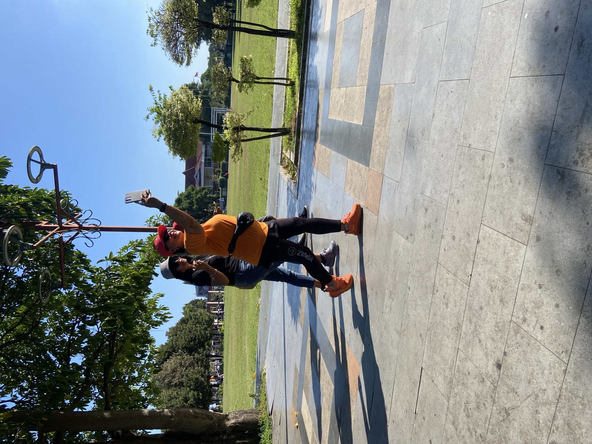 Two women are walking on a paved walkway in front of a grassy field under a clear blue sky. The woman in the foreground, on the right, is wearing an orange t-shirt, black leggings with "Zumba" written on the leg, and bright orange athletic shoes. She is holding up a smartphone in her right hand, appearing to take a selfie as she walks. She also has a black fanny pack around her waist. She is wearing a red baseball cap. The woman behind her, on the left, is wearing a black t-shirt, blue jeans, and grey athletic shoes. She is wearing a grey baseball cap and sunglasses. She is looking towards the woman in front of her and has her left hand raised with two fingers extended. A large tree with green leaves is to the left of the frame, casting shadows on the walkway. In the background, there is a large green field, several small trees with light green foliage, and a building with a red roof. The walkway is paved with large, rectangular tiles of varying colors, including grey, beige, and pinkish-brown. This scene appears to be in a public park or outdoor recreational area in Kecamatan Pati, Indonesia.