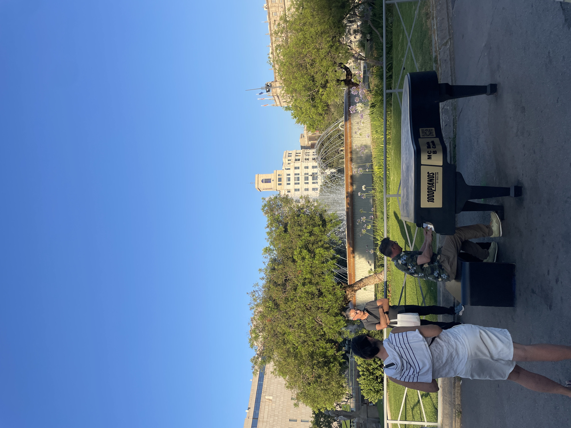 In a sunlit urban park, several people are gathered around a black piano-shaped bench. The scene is set against a backdrop of distinct architecture, including a tall, cream-colored building with numerous windows and a larger, imposing stone building with arched windows and flags. A fountain with water arching upwards is visible in the mid-ground, surrounded by lush green trees and flowering plants. The sky is a clear, vibrant blue, suggesting a pleasant daytime.
One person, wearing glasses and a dark t-shirt, stands near the piano bench, looking towards the fountain. Another individual, in a floral shirt and khaki pants, is seated on the piano bench, engrossed in a mobile phone. A third person, wearing a white shirt with dark stripes and white shorts, stands with their back to the camera, facing away from the bench. The piano bench itself has text on it, including "1000PIANOS" and "BY WOWDM". There are also what appear to be QR codes and other symbols on the bench. The overall atmosphere seems relaxed and casual, typical of a public park on a bright day. The presence of the piano bench suggests a public art installation or an event related to music.