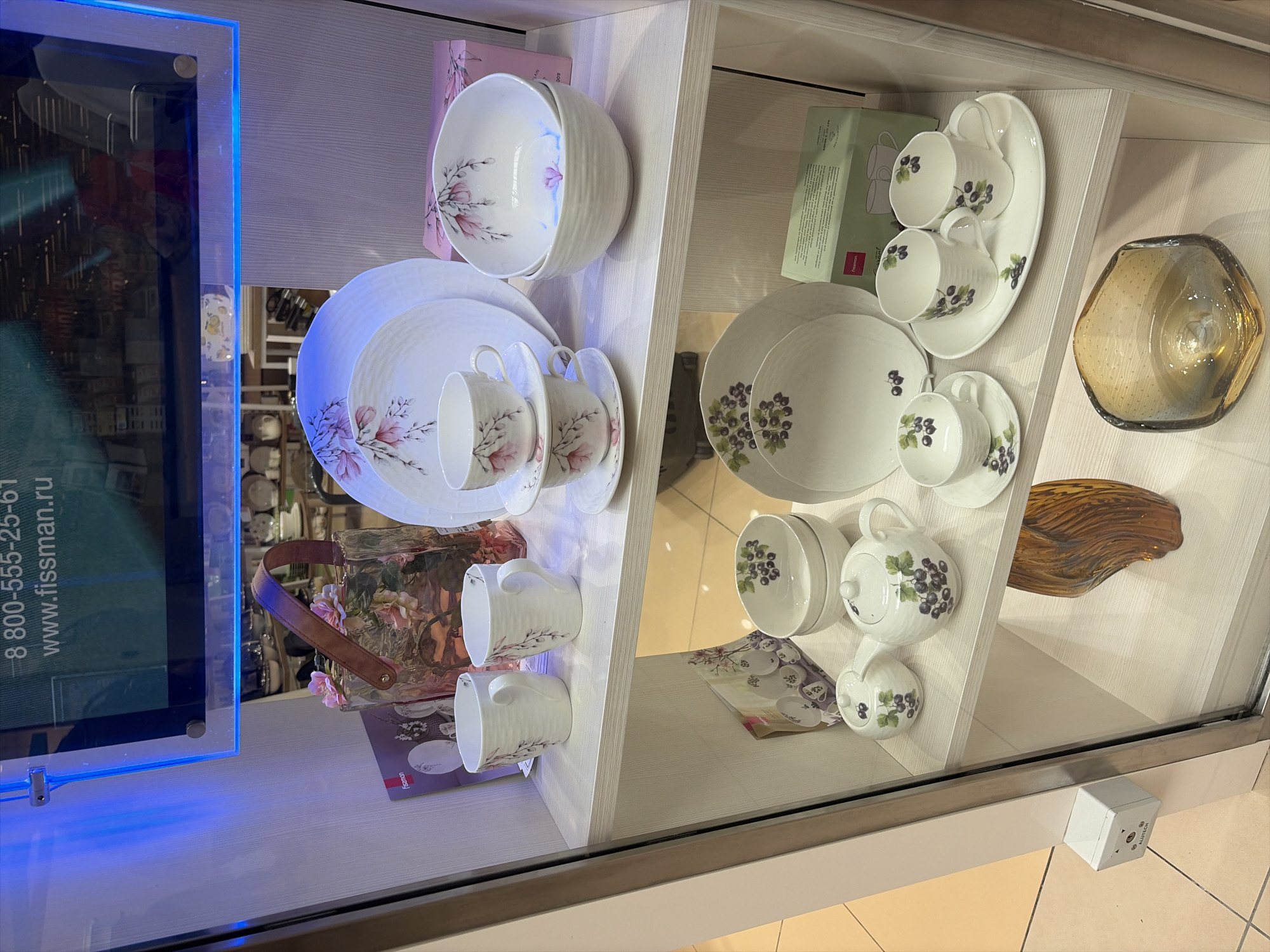 The image depicts a display case in a store, likely a home goods or kitchenware retailer, located in Balashikha, Russia. The display case is illuminated, highlighting various sets of white ceramic tableware.
On the top shelf, there are several white ceramic cups with delicate floral patterns, and stacked plates. A decorative glass container with a leather handle and floral accents is also present on this shelf.
The middle shelf features more white ceramic tableware, including cups, saucers, and bowls, also adorned with floral motifs. A pink box with floral designs is visible to the right.
The bottom shelf displays a teapot and sugar bowl, both decorated with a pattern of dark berries and leaves. A stack of plates and cups with the same berry motif are also visible, alongside a brochure. Further to the right on this shelf is a amber-colored glass decorative bowl.
A small white electrical device is mounted on the exterior of the display case on the left side. The background shows reflections of store lighting and shelves stocked with additional merchandise, suggesting a retail environment. The overall impression is a well-organized and aesthetically pleasing presentation of tableware.