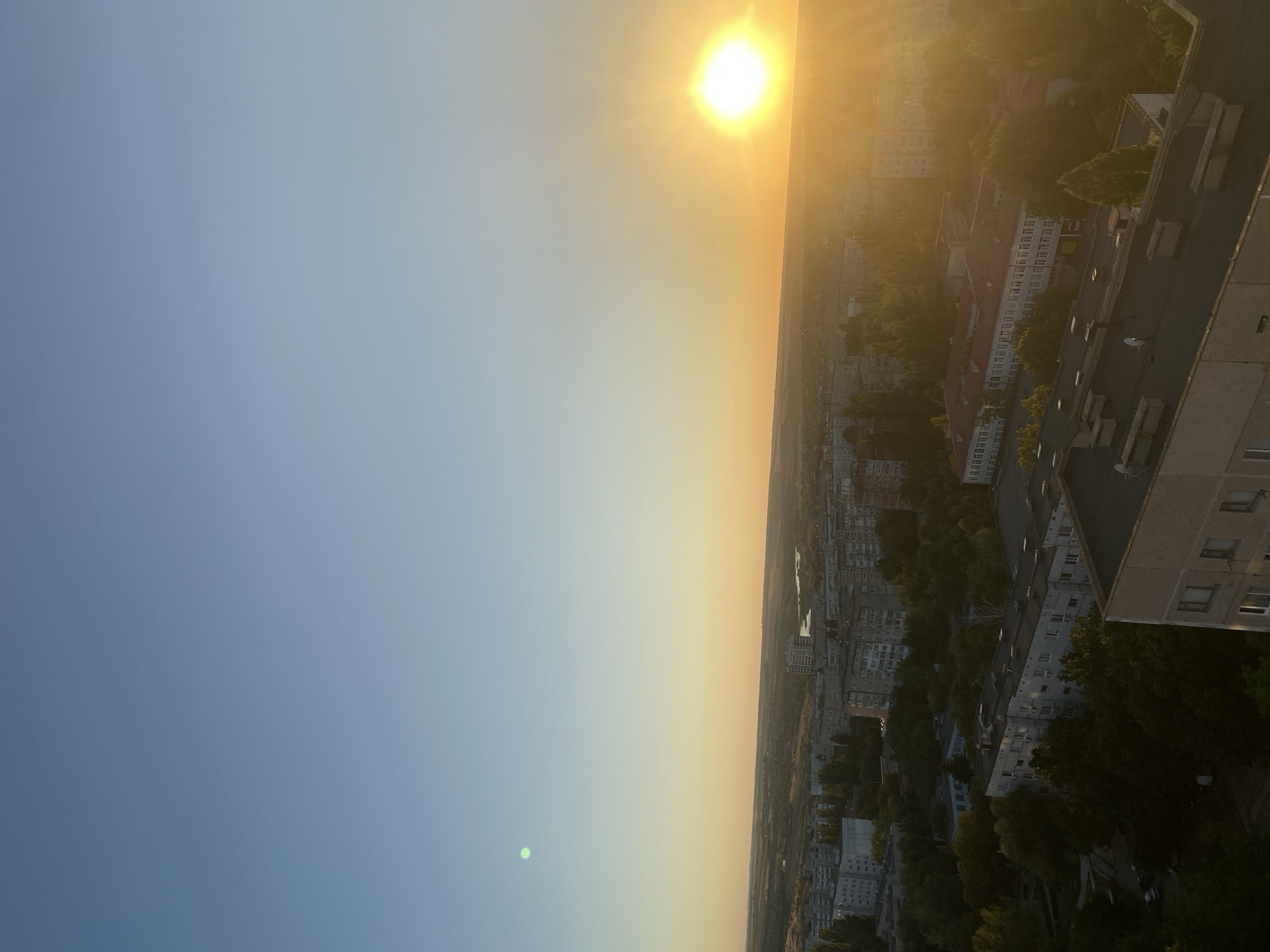 This image depicts an aerial view of the city of Pivdennoukrainsk, Ukraine, during a sunset. The sun is prominently visible on the right side of the frame, casting a warm, golden light over the cityscape and horizon. The sky transitions from a clear blue at the top to a gradient of orange and yellow near the horizon.
The scene is dominated by residential buildings, characterized by their multi-story architecture, typical of Soviet-era urban development. Many of these buildings are apartment blocks with visible windows and balconies. Dense foliage, primarily trees, is interspersed throughout the urban landscape, creating pockets of green amidst the concrete structures. A body of water, likely a river or reservoir, is visible in the distance, reflecting the warm hues of the sunset.
There are no people or animals clearly visible in the image. The focus is on the urban environment and the natural phenomenon of the sunset. The overall impression is one of a quiet, residential area at the end of the day, with the natural beauty of the sunset enhancing the scene.