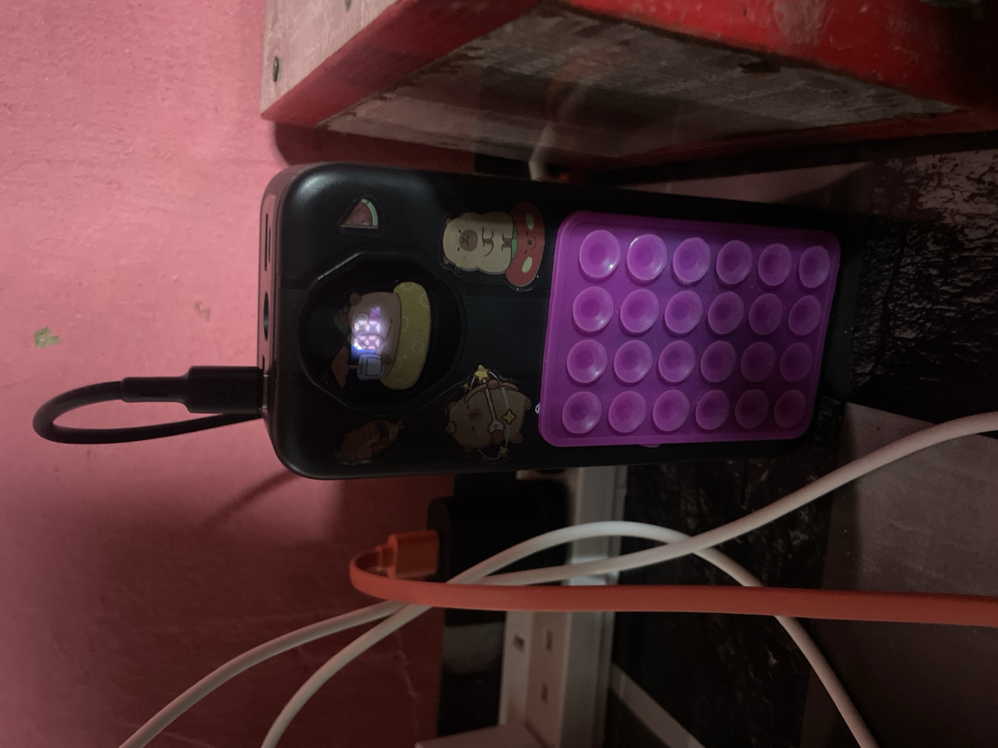 The image shows a black portable charger with several stickers on it, including cartoon characters and a slice of watermelon. A purple pop-it toy is attached to the bottom of the charger. A black cable is plugged into the top of the charger, and an orange cable is plugged into a white power strip nearby. The charger and cables are on a dark surface, possibly a table, with a pink wall in the background. The scene appears to be indoors. There are no people or animals visible in the image. The location is specified as Jalingo, Nigeria, but there are no visual cues in the image to confirm this location.
