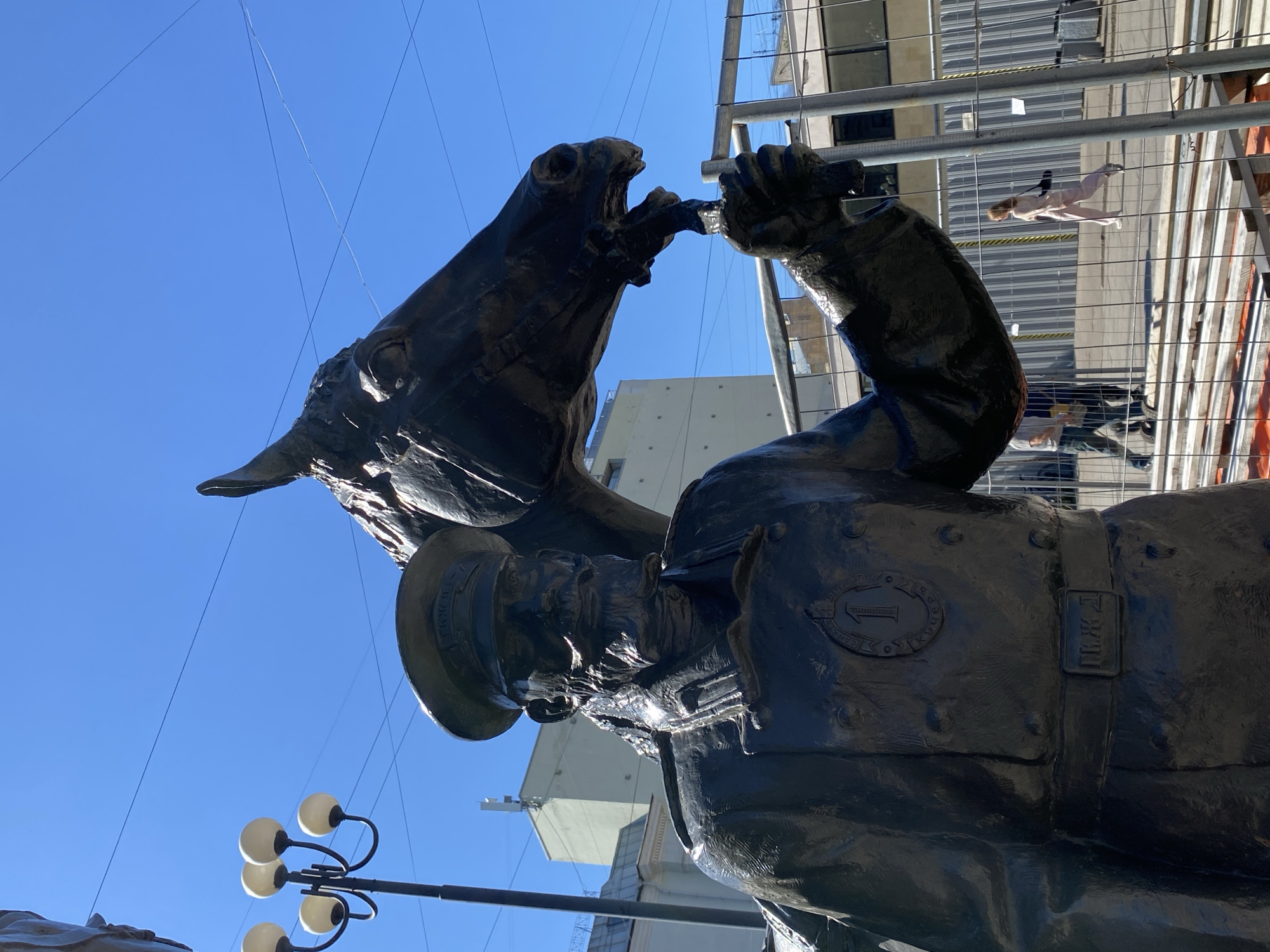 The image displays a large, dark bronze statue of a man and a horse, viewed from a low angle against a clear blue sky. The man, depicted as a uniformed figure with a peaked cap, is offering a drink to the horse from a cup or goblet held in his right hand. The horse has its head raised and its mouth open, appearing to drink from the vessel.
In the background, modern urban architecture is visible, including buildings with glass facades and metal construction scaffolding or fencing. Through the fencing, a faint outline of a mannequin or person in light-colored attire can be seen. A street light featuring multiple spherical lamps is present in the lower left portion of the image.
The scene is brightly lit by strong daylight, casting defined shadows and highlights on the statue, indicative of sunny weather. Visible text on the statue includes Cyrillic letters on the man's cap, which appear to be "Т.К.Ж.Т." or "Г.К.Ж.Т.". On his uniform, an oval emblem contains the number "1" and is surrounded by faint Cyrillic text, possibly "ПЕРВЫЙ" (First). Below this, a rectangular buckle or emblem on his belt clearly shows the Cyrillic letters "П.К.Ж.Д.". The statue captures a moment of interaction and care between the man and the horse, set against a bustling urban backdrop.