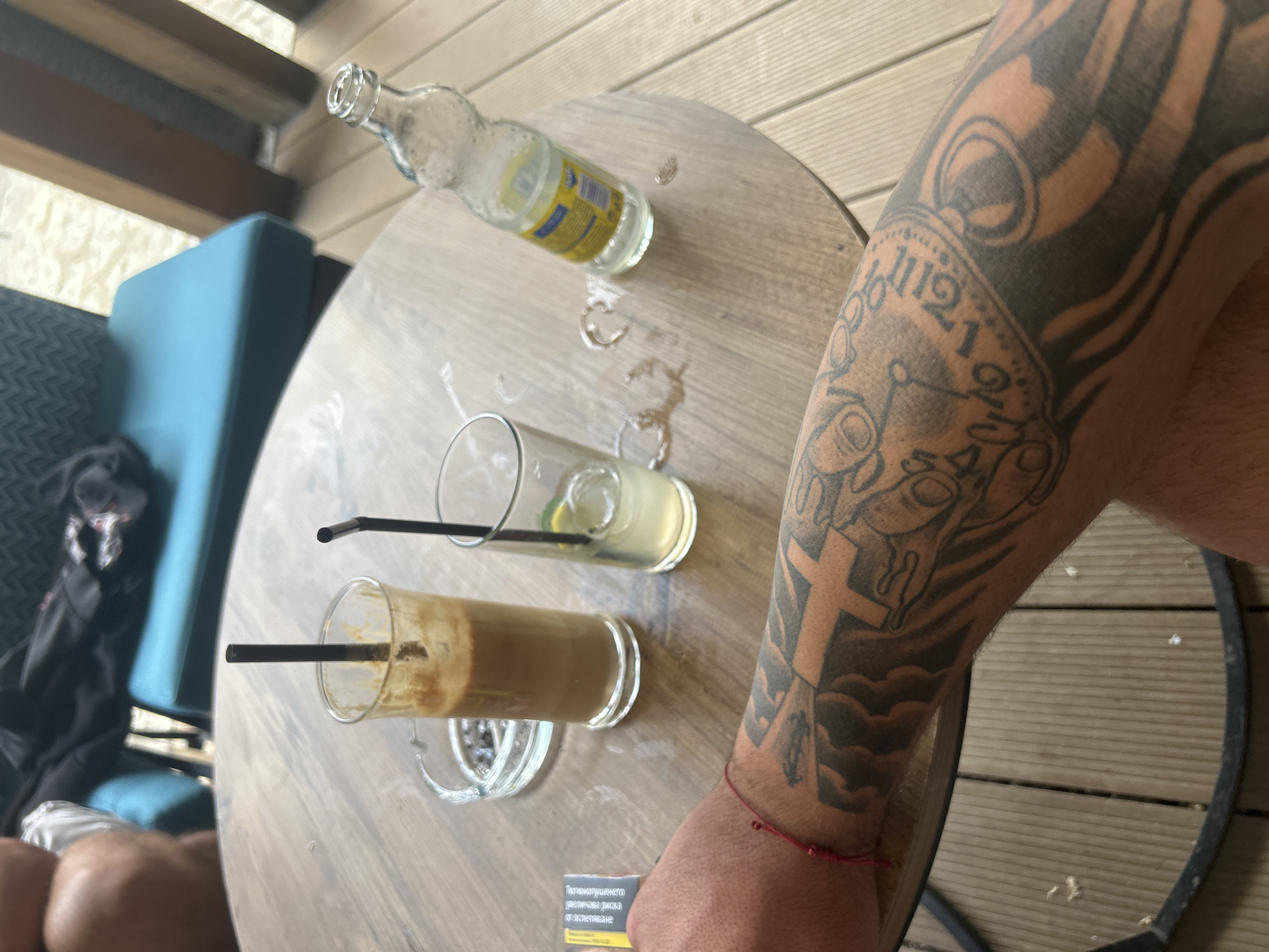 The image presents a close-up, high-angle view of a person's arm and a wooden table, likely in an outdoor or semi-outdoor setting such as a balcony or cafe in Razlog, Bulgaria. The primary subject is a heavily tattooed arm, featuring a sleeve design in grey and black. The tattoo includes a prominent pocket watch or clock face with Roman numerals and Arabic numbers like "12," "1," "2," and "11," along with religious symbols such as crosses, cloud formations, and other abstract patterns. A thin red string bracelet is visible on the person's wrist.
On the light wooden table, there are two used drinking glasses with black straws. One glass contains the remnants of a layered iced coffee. The other glass holds a small amount of clear liquid, ice, and a lime wedge, indicating a finished lemonade or similar cold drink. An empty clear glass bottle with a yellow label rests on its side, and several water rings and condensation marks are visible on the tabletop. A clear glass ashtray, currently empty, is also on the table.
In the background, a part of a blue cushioned chair is visible, with a black bag placed upon it. The flooring appears to be light-colored wooden planks. A white and orange cigarette warning label is partially visible on the table, with text in Bulgarian: "ПУШЕНЕТО причинява рак и смъртоносни белодробни заболявания." (SMOKING causes cancer and fatal lung diseases.) and a helpline number "0800 900 35". The overall scene suggests a casual, relaxed moment during the daytime.
