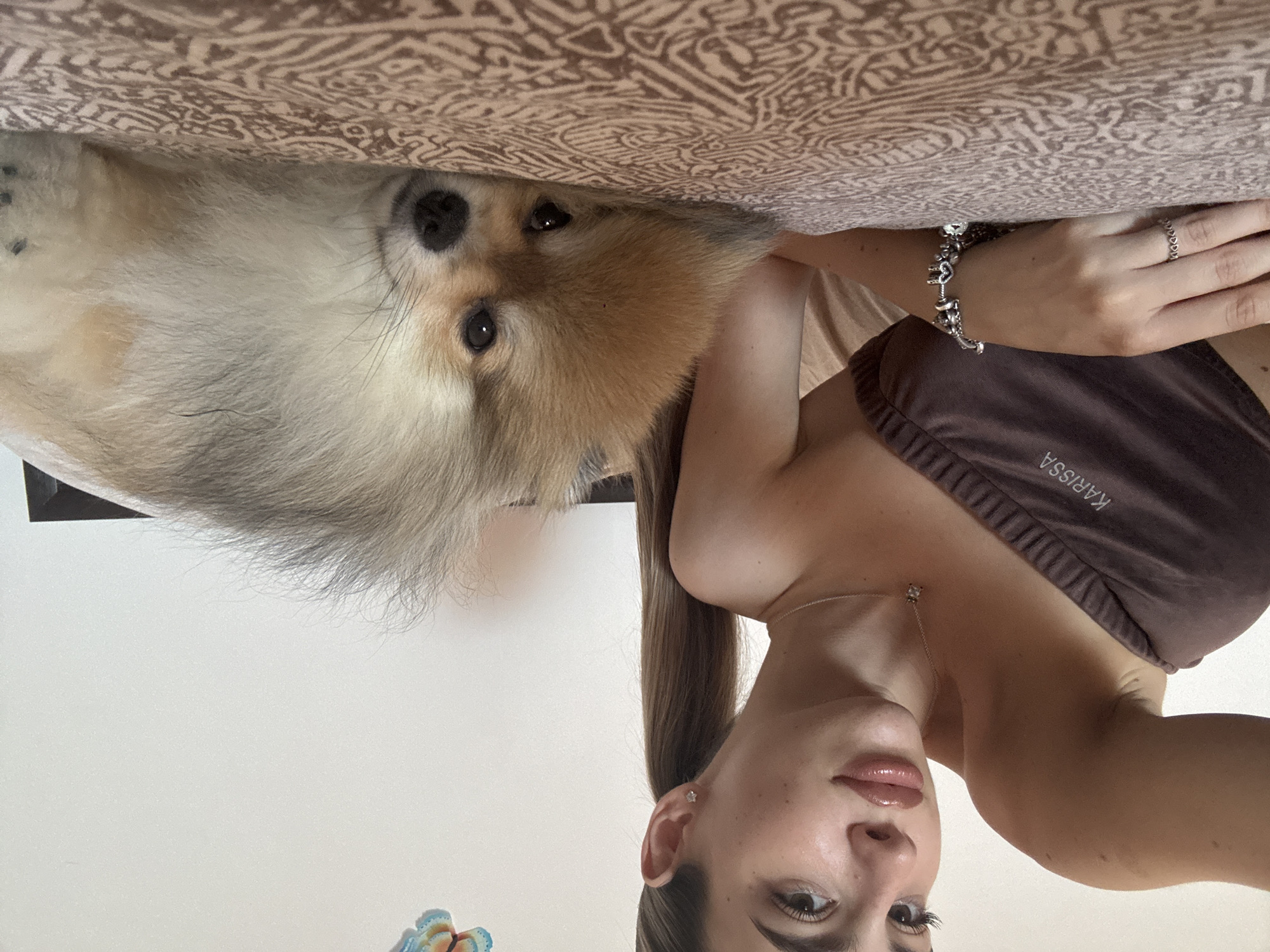 A young woman with long, light brown hair pulled into a ponytail lies on a bed next to a small, fluffy Pomeranian dog. The woman is wearing a brown strapless top with the word "KARISSA" printed on it in white lettering. She is wearing a necklace with a small pendant and a bracelet adorned with heart-shaped charms, as well as a ring on her left ring finger. The dog is lying on its side, facing the camera, with its head resting on the bed. The bedspread has a patterned design. The background appears to be a plain white wall, with a decorative butterfly sticker visible in the upper right corner. The setting is likely an indoor bedroom. No specific elements in the image directly indicate the location as Nesebar, Bulgaria.