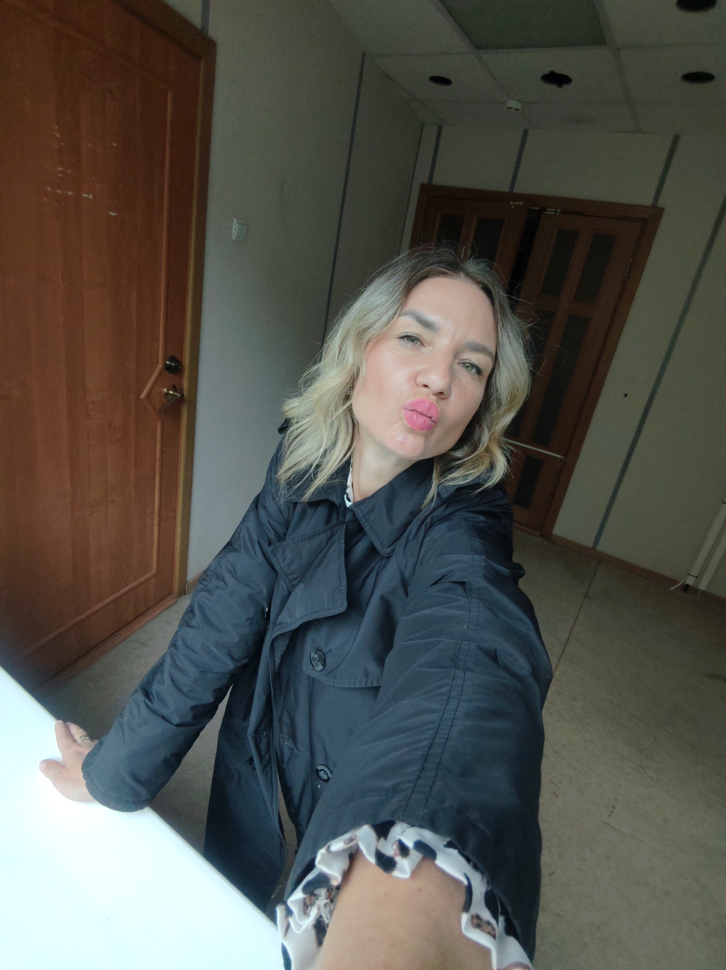 A woman with blonde hair, wearing a black jacket, is posing for a selfie. She is puckering her lips and looking at the camera. The background includes two wooden doors and beige walls. The location appears to be an indoor space with a drop ceiling.
