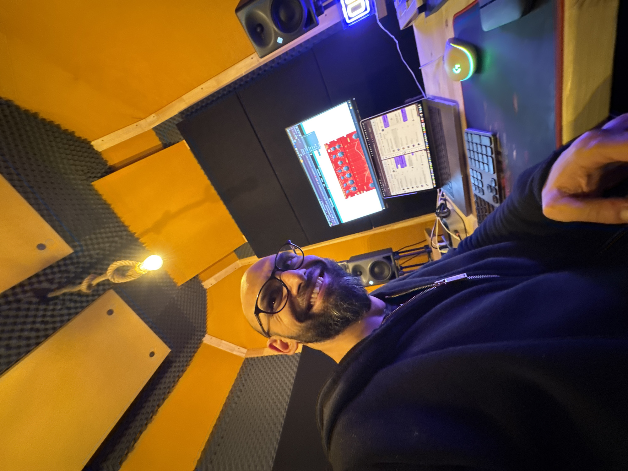 The image depicts a man with glasses and a beard in what appears to be a recording studio, likely in Plovdiv, Bulgaria. The environment features soundproofing material on the walls, indicative of acoustic treatment. There is a computer setup with dual monitors, a keyboard, and a mouse with a light-up feature. One monitor displays audio editing software. Speakers are visible above the monitors, suggesting a setup for audio production or mixing. A lightbulb hangs from the ceiling. The man is wearing a black zippered jacket and is smiling, looking at the camera.