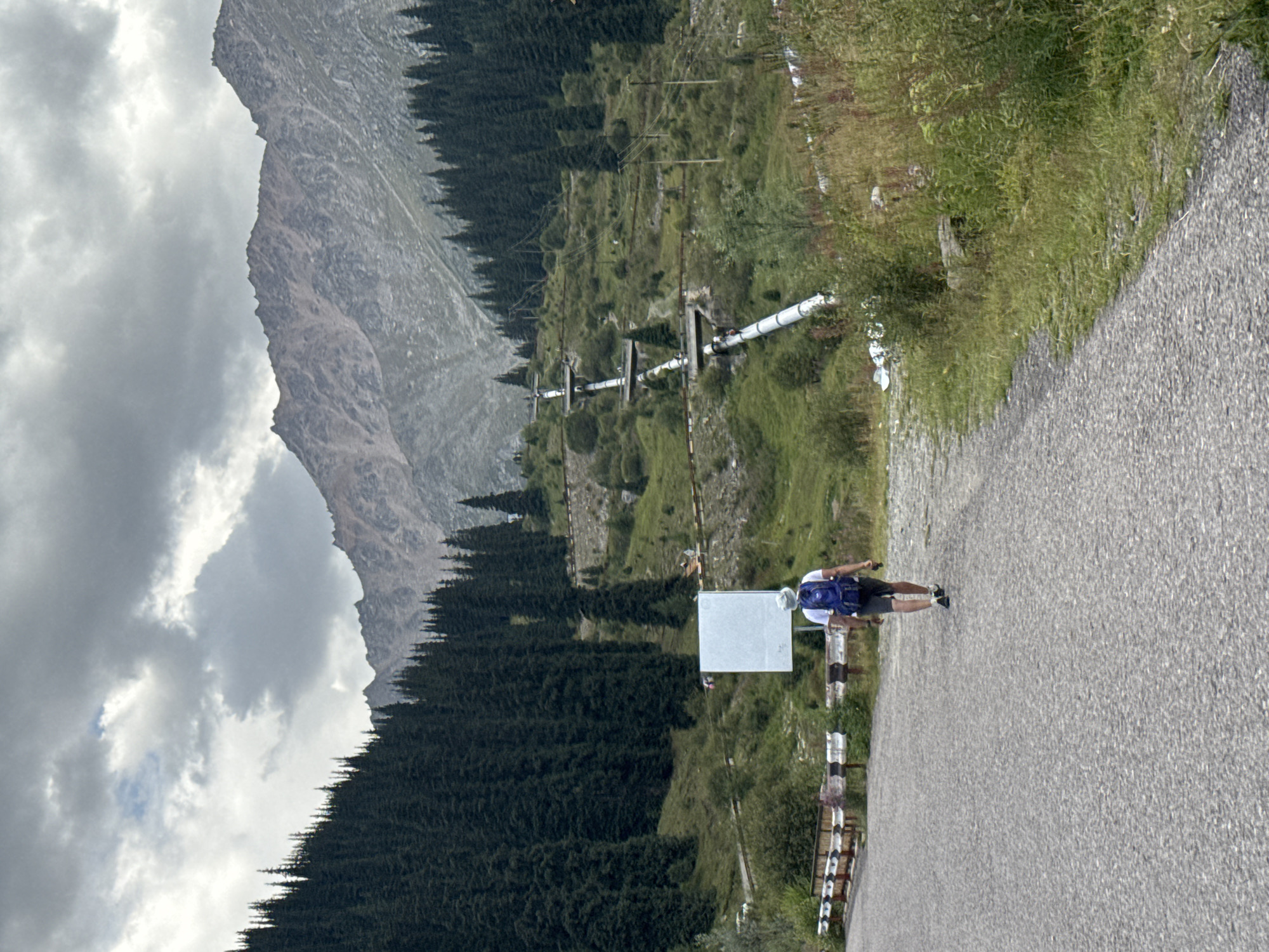 A solitary person wearing a white hat, white t-shirt, dark shorts, and a blue backpack walks away from the camera on a paved road. To the left of the road is a metal guardrail with white and black stripes. Further up the slope to the right is a large, blank white signpost. The road leads towards a mountain landscape with a dense forest of dark green coniferous trees covering the lower slopes. A prominent white pipe structure snakes its way up the mountain, supported by pylons. The upper slopes of the mountain are rocky and barren, with a cloudy, grey sky overhead. The scene appears to be in a mountainous area near Almaty, Kazakhstan, likely in a region with infrastructure for water or energy transport.